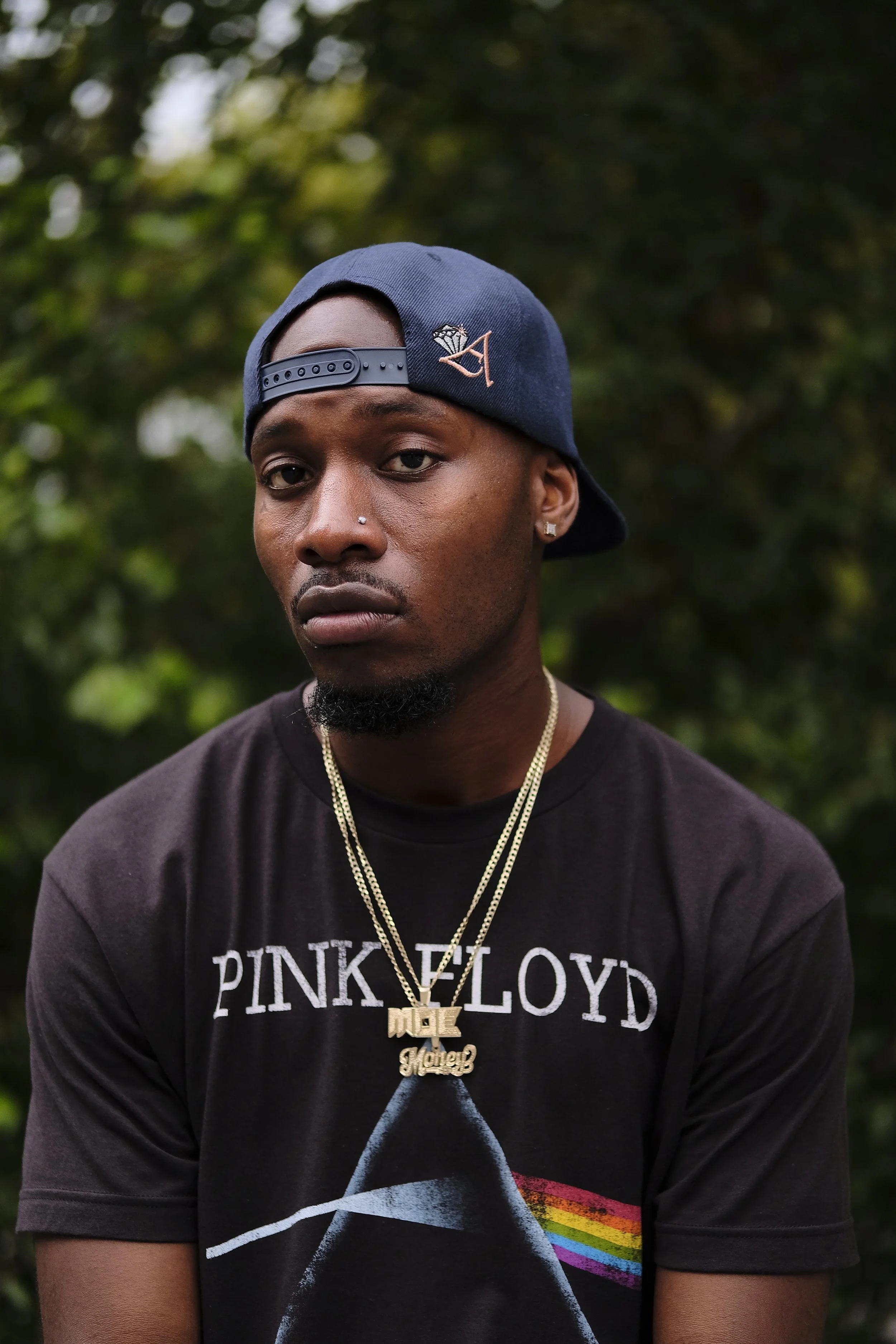 A young man wearing a blue baseball cap backwards, a black Pink Floyd t-shirt, gold chains, and diamond earrings, standing outdoors with a blurred green leafy background.