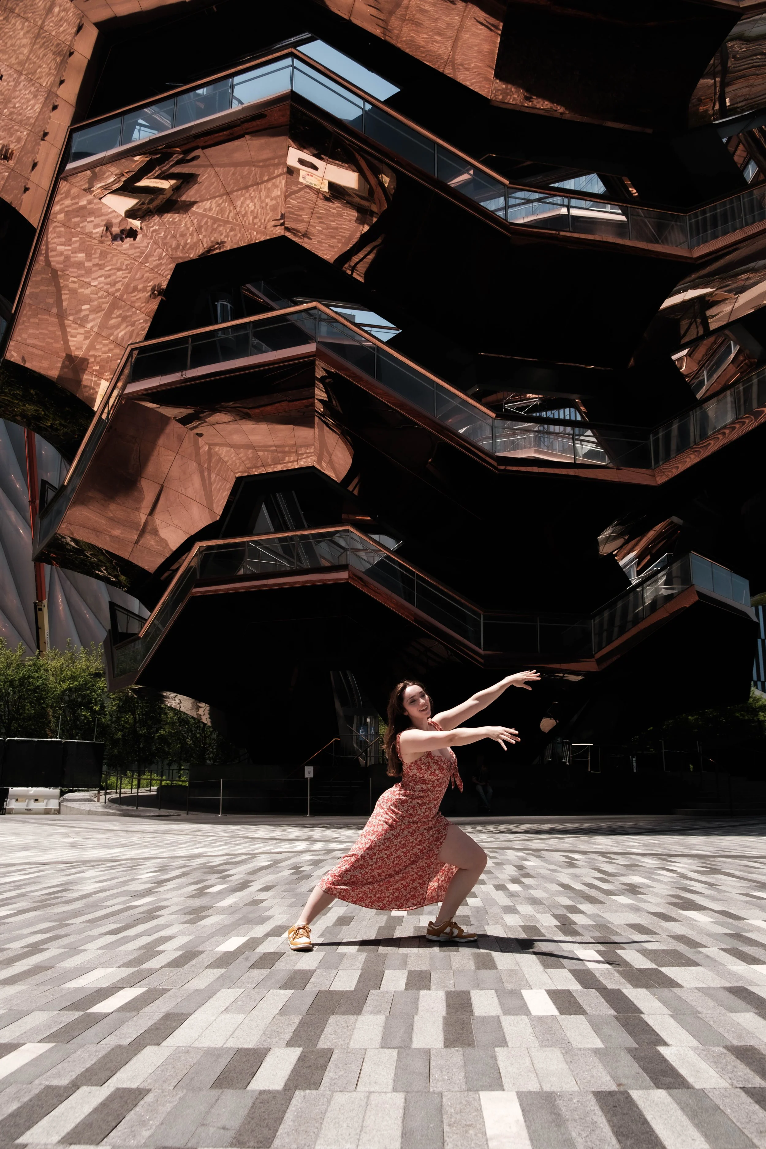 A woman in a red floral dress with white sandals poses in a wide stance on a checkered pavement in front of a modern building with glass balconies and reflective copper-colored panels, during daytime.