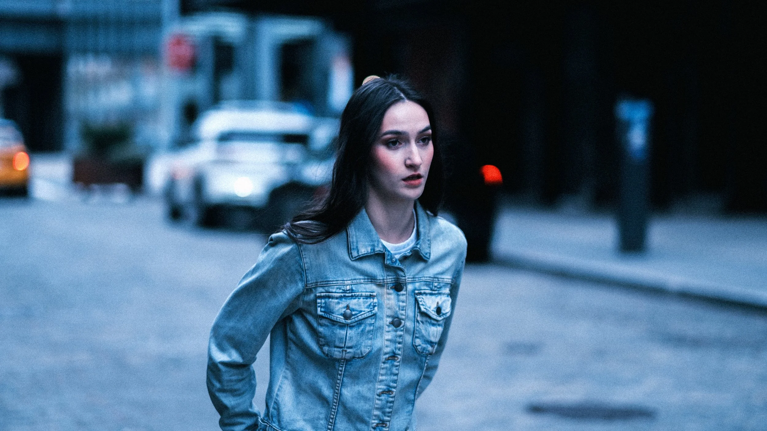 A young woman with dark hair, wearing a denim jacket, walking on a city street during evening hours, with blurred cars and city buildings in the background.