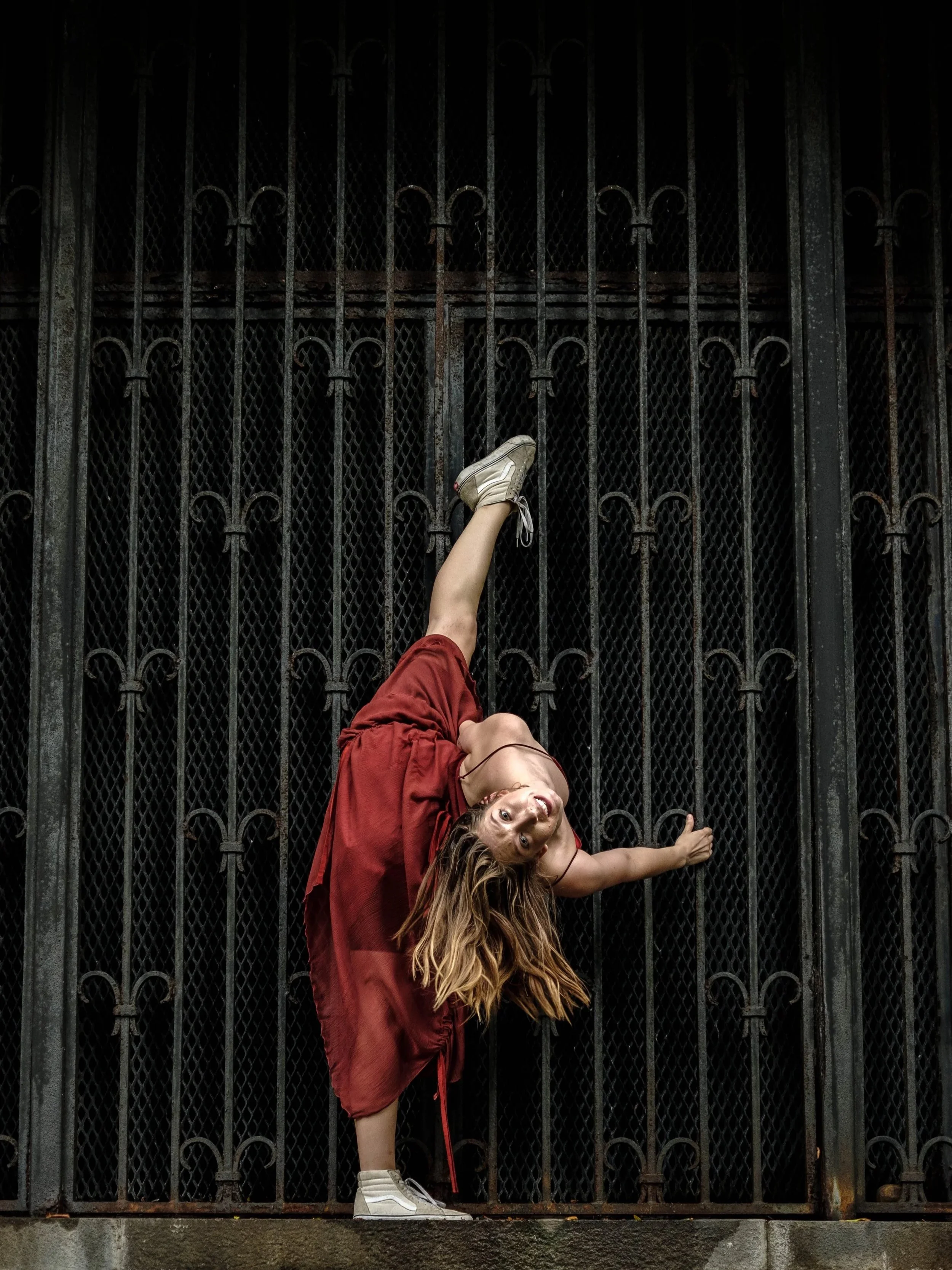 A person in a red dress and white sneakers is performing a handstand against a black iron gate, with their head near the ground and their legs extended upward, in an urban setting.