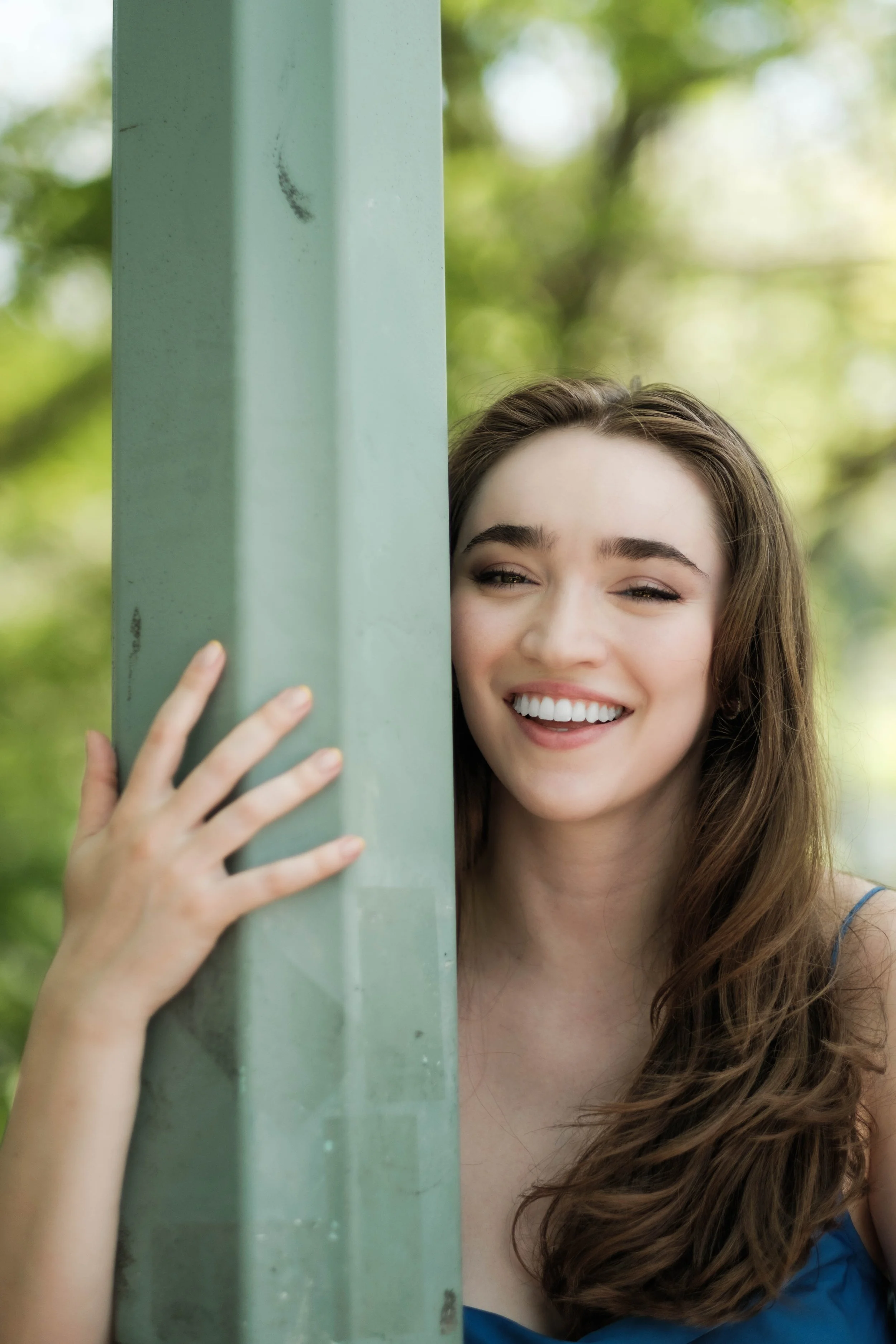 Close-up of a woman with long brown hair and a bright smile, standing behind a vertical green pole, with a background of green blurred trees.