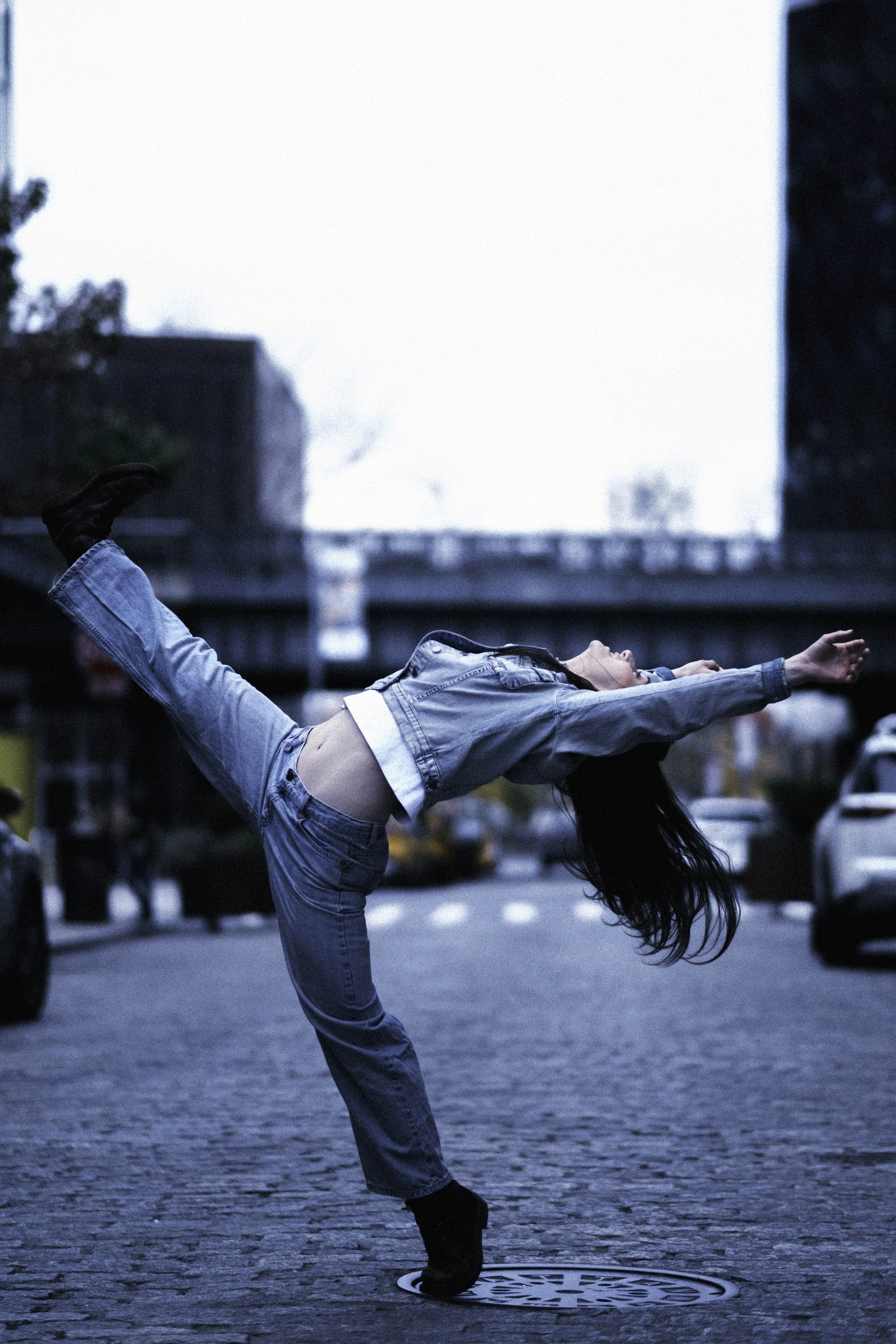 A woman performing a handstand on a city street in the early evening, wearing a denim jacket and jeans, with buildings and cars in the background.