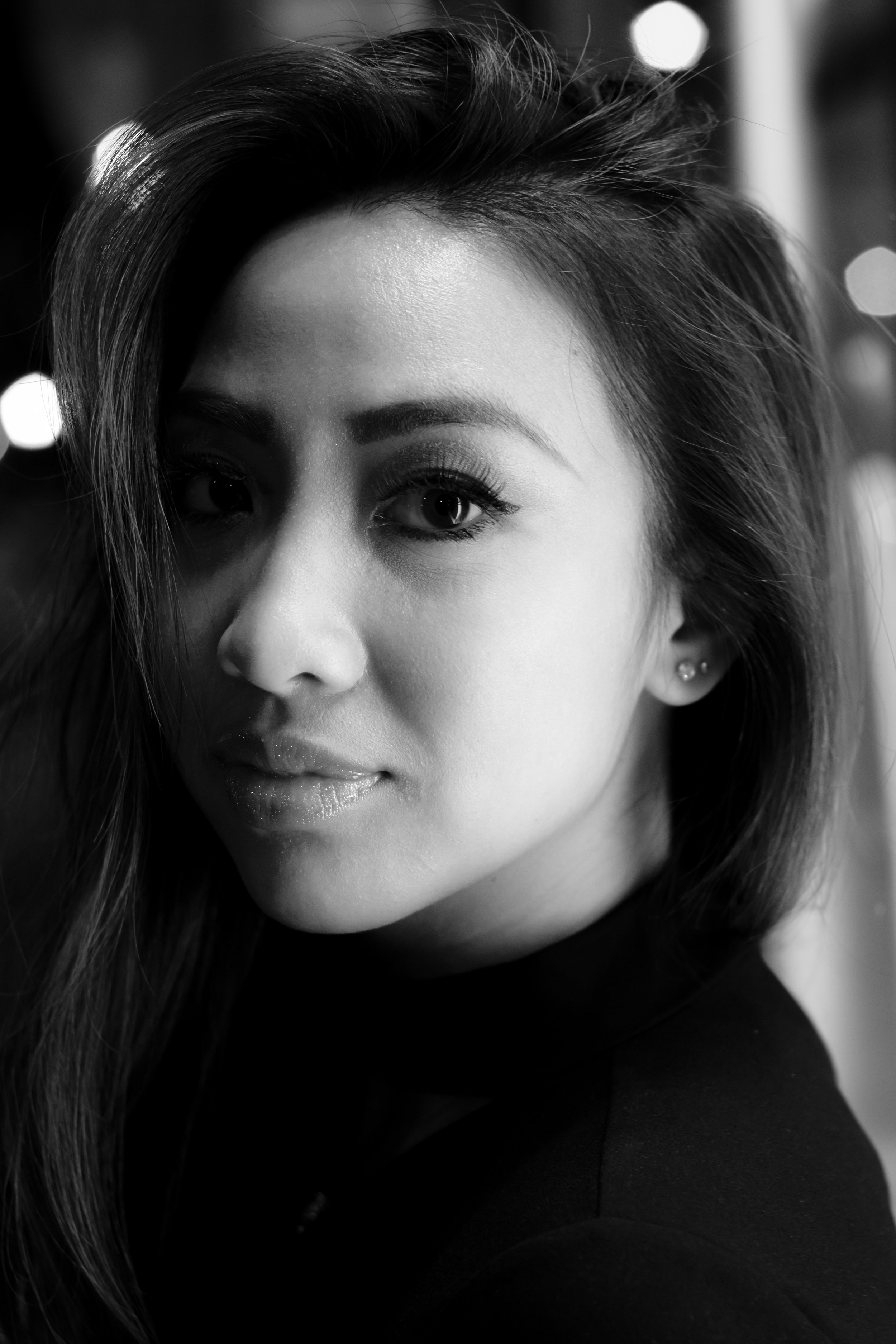 Black and white close-up portrait of a woman with long hair, partially covering one eye, looking directly at the camera, with a blurred bokeh background.