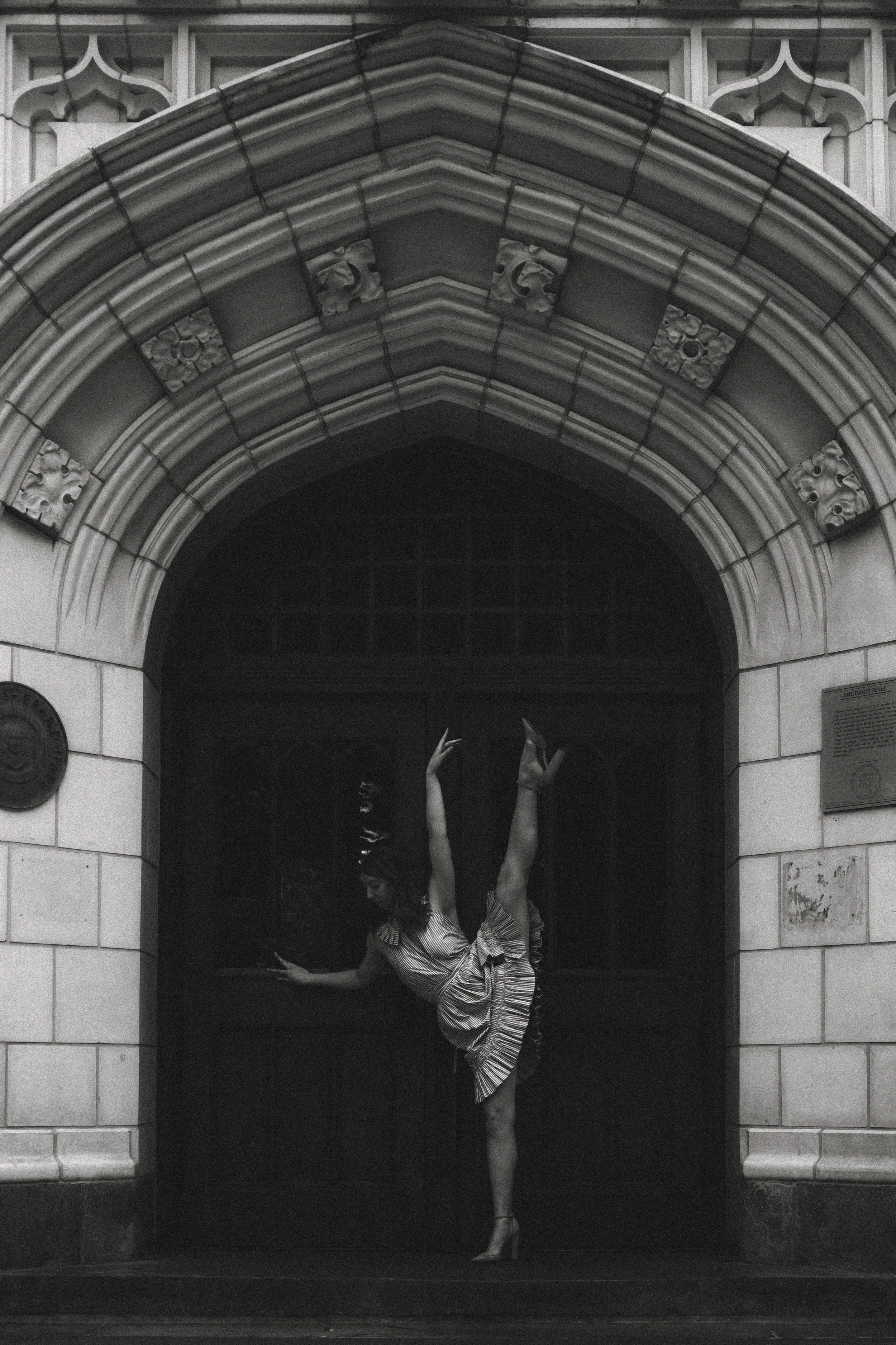 A woman dancing in a theatrical pose under an ornate stone archway entrance to a building, wearing a striped dress and high heels.