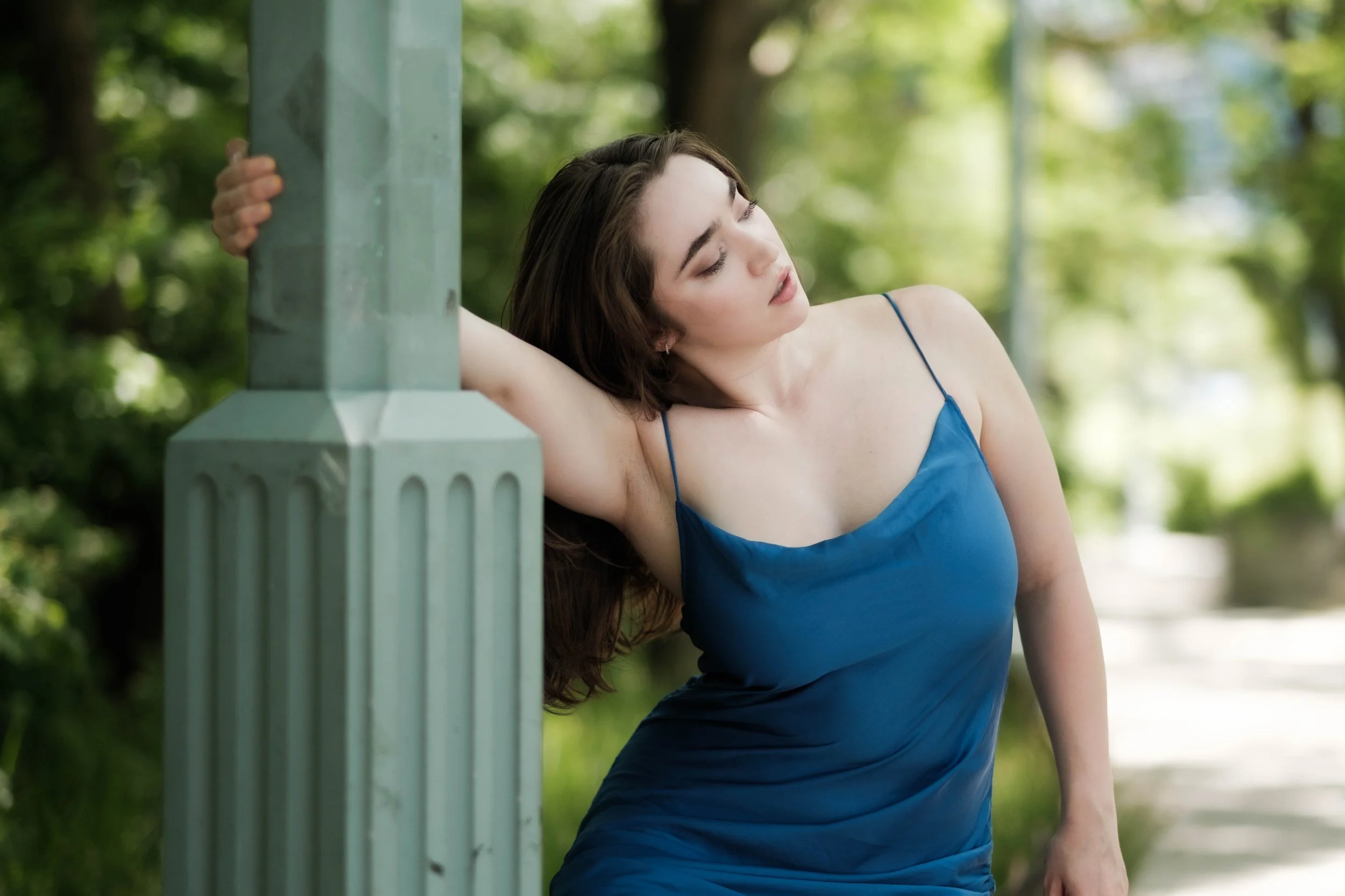 A young woman in a blue dress leaning against a green park bench with her eyes closed, surrounded by green trees and sunlight.