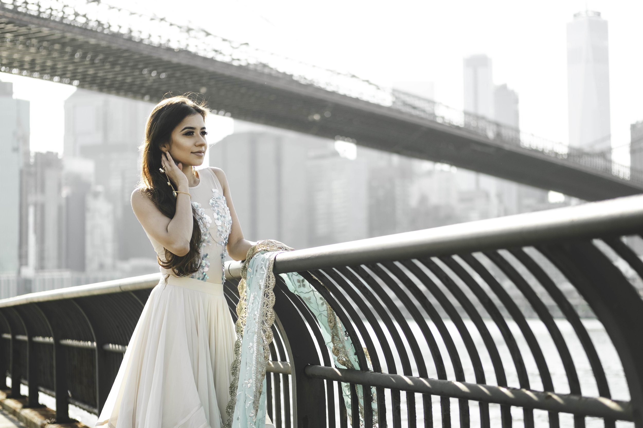 A woman with long dark hair, wearing a white dress with floral embellishments, stands by a curved bridge railing in an urban setting. The background features a city skyline with tall buildings and a bridge overhead.