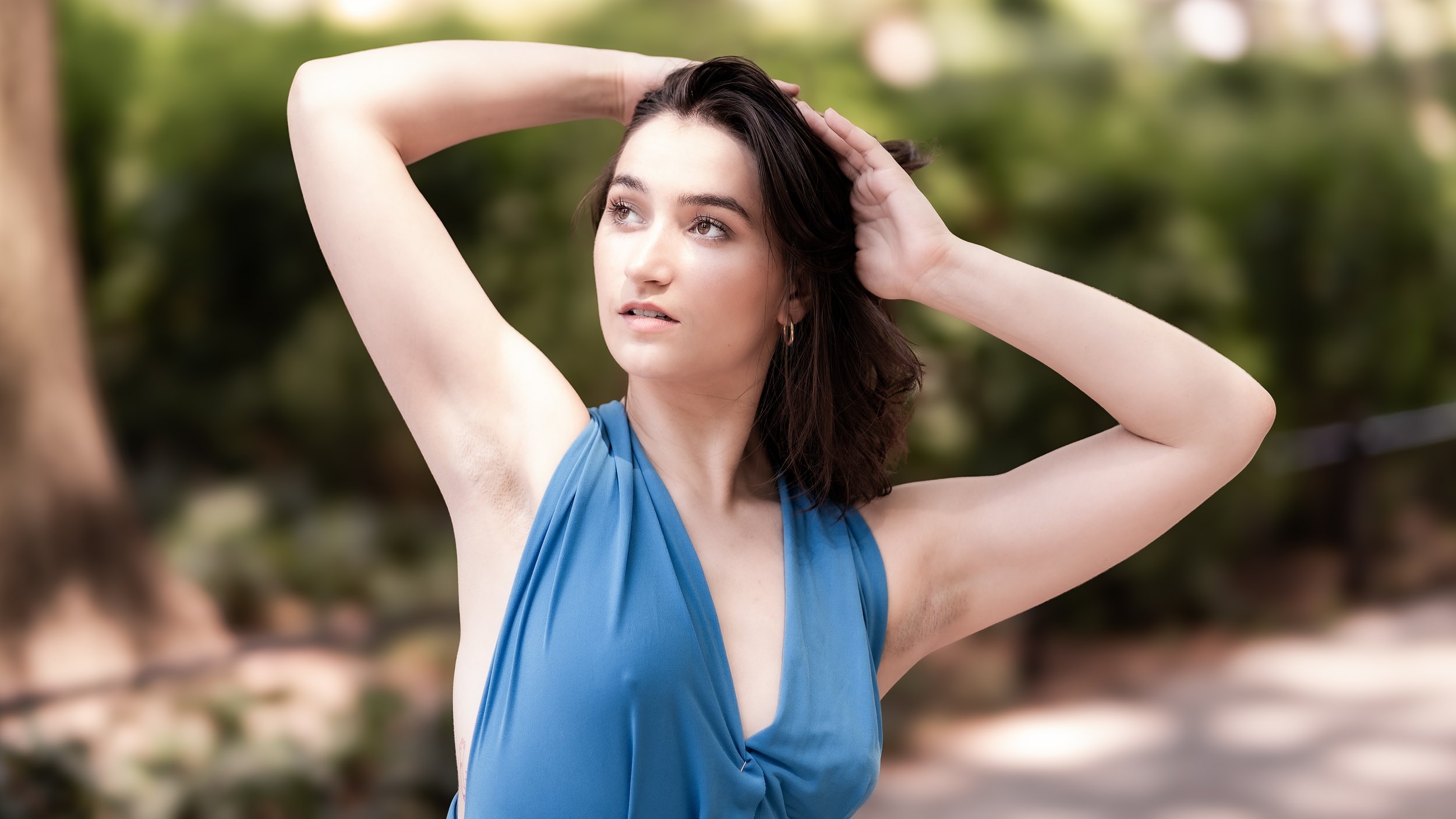 A woman with dark hair and light skin, wearing a blue sleeveless top, outdoors in a park, with a blurred green background, looking off to the side with her hands in her hair.