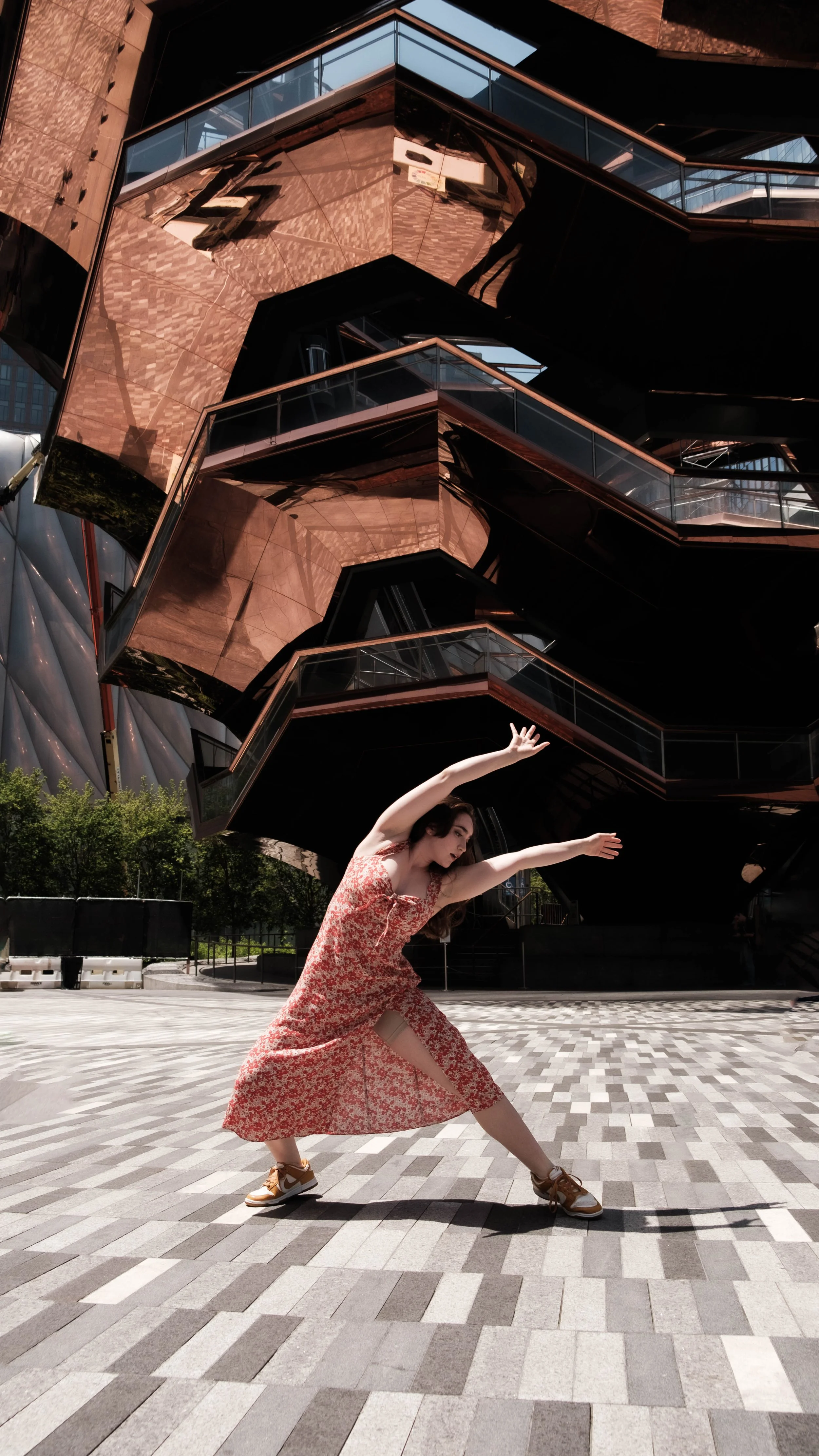 A young woman in a red floral dress and sneakers in a dance pose outdoors, with a modern building featuring geometric design and reflective copper panels in the background.