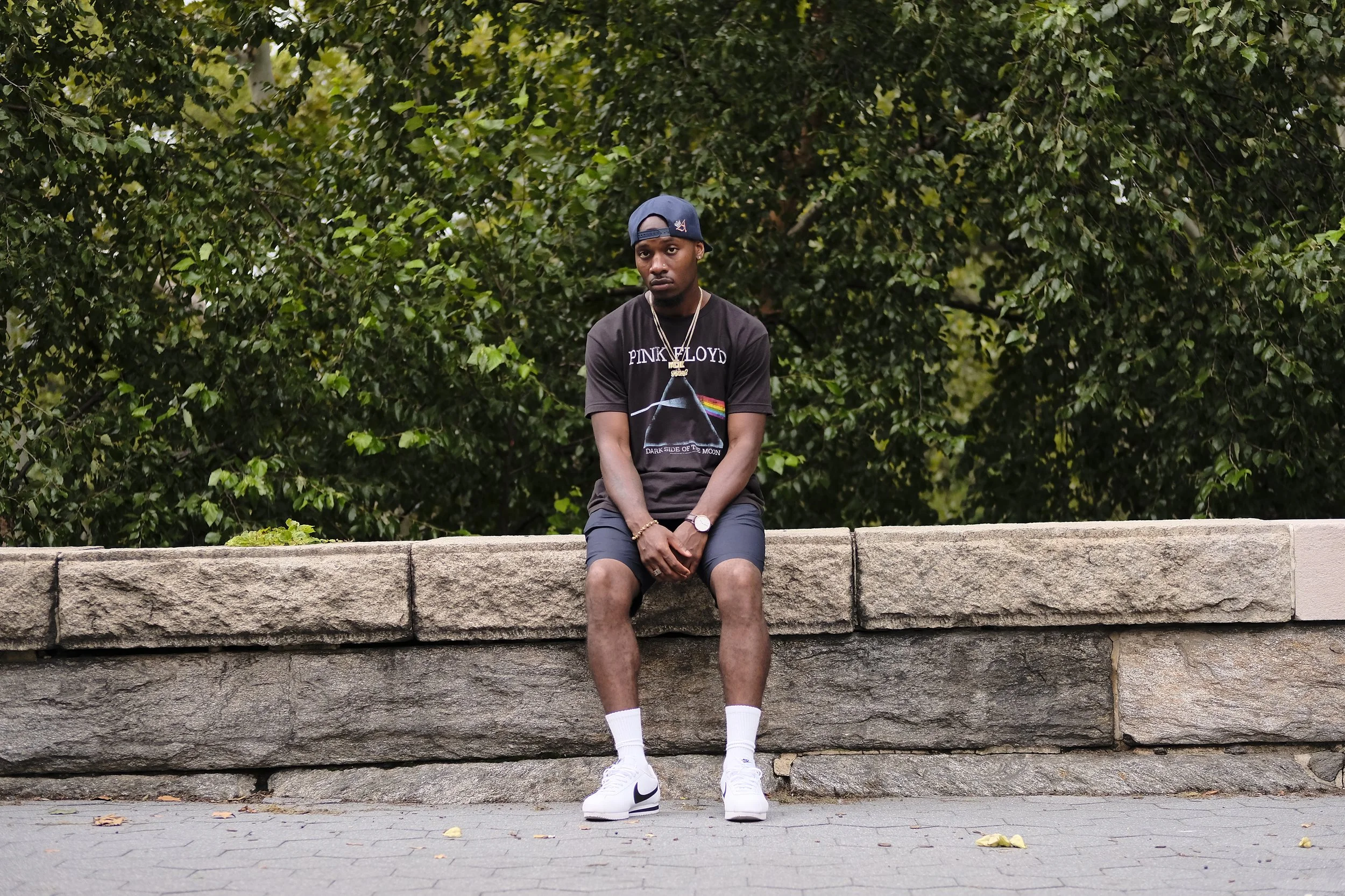 Young man sitting on a stone ledge in front of a green leafy background, wearing a black T-shirt, shorts, white sneakers, a cap backwards, and jewelry.