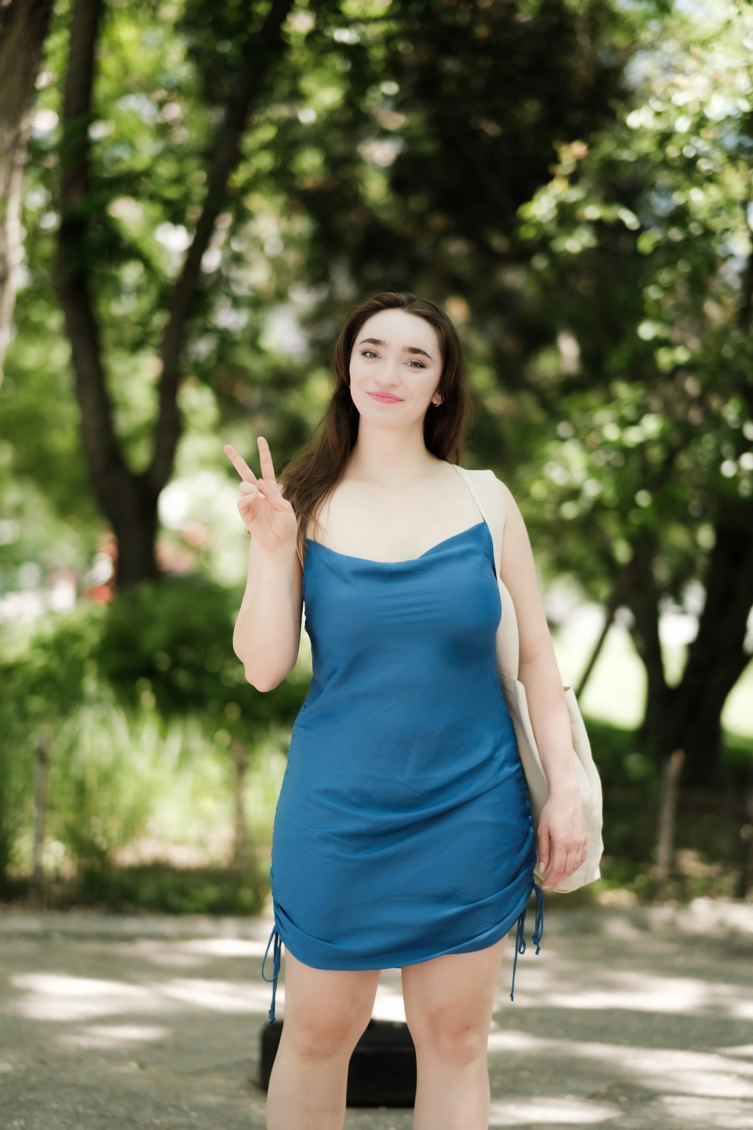 A young woman with brown hair in a blue dress making a peace sign with her right hand while standing outdoors with trees in the background.