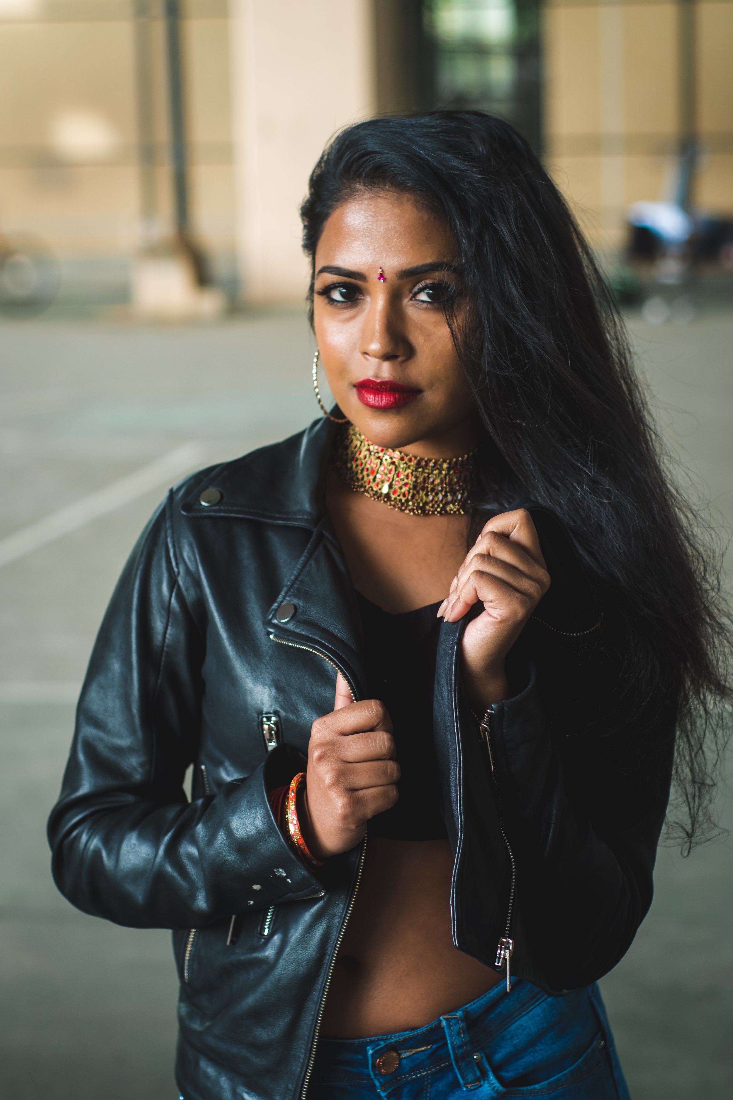 A woman with dark hair, wearing a black leather jacket, red lipstick, colorful jewelry, and hoop earrings, standing outdoors in front of a blurred background.