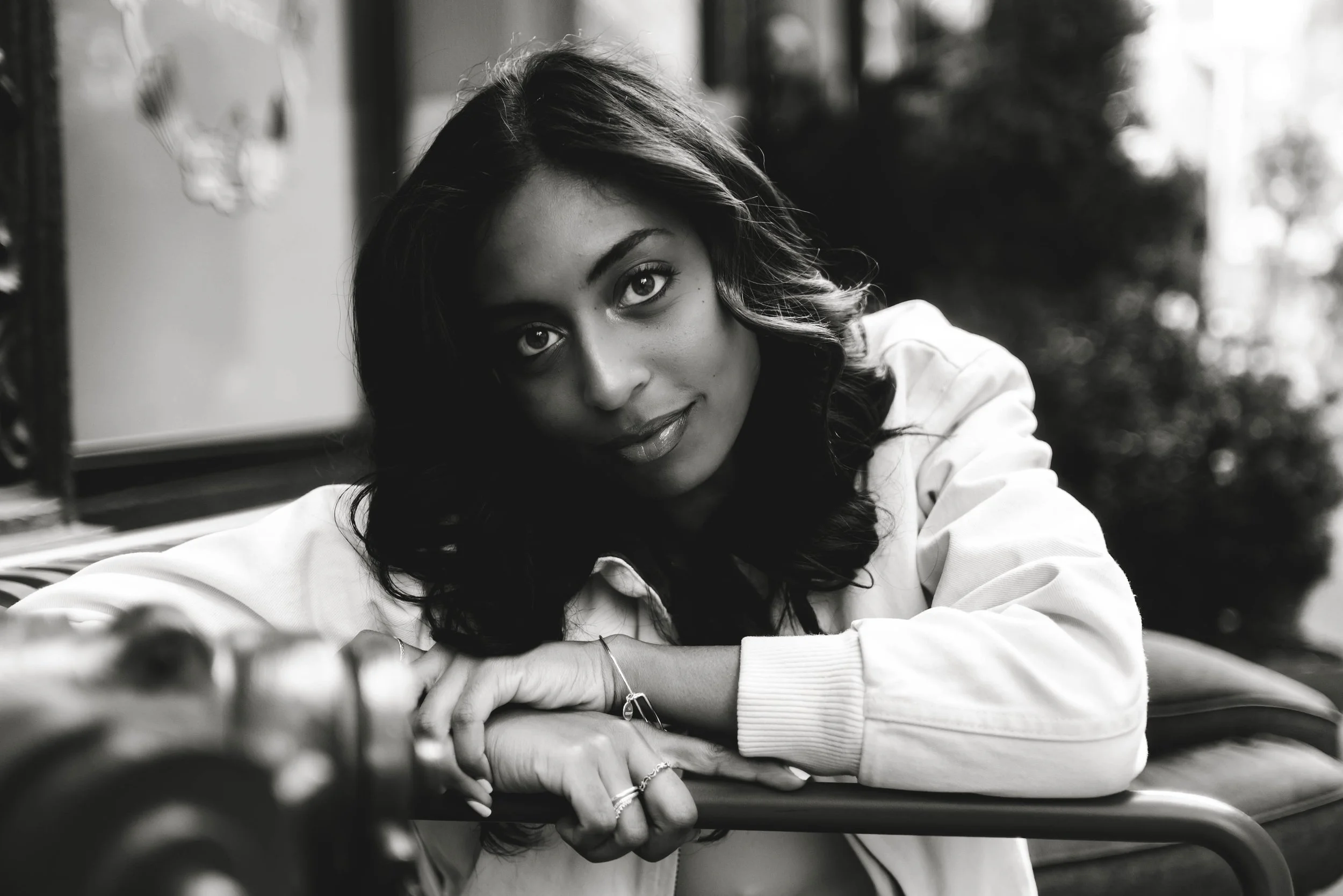 Black and white close-up of a woman with long hair, leaning on her arms, looking at the camera with a soft expression, outdoors with blurred trees in the background.