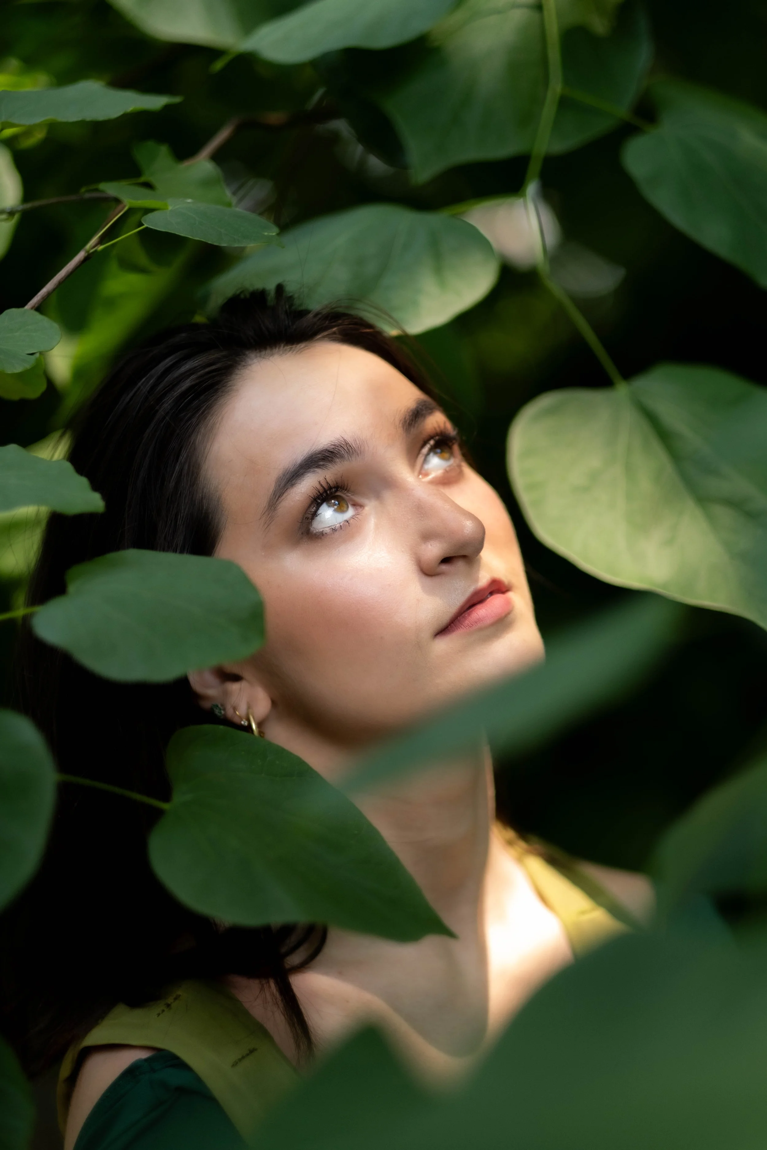 Young woman with dark hair and brown eyes looking upward surrounded by large green leaves.