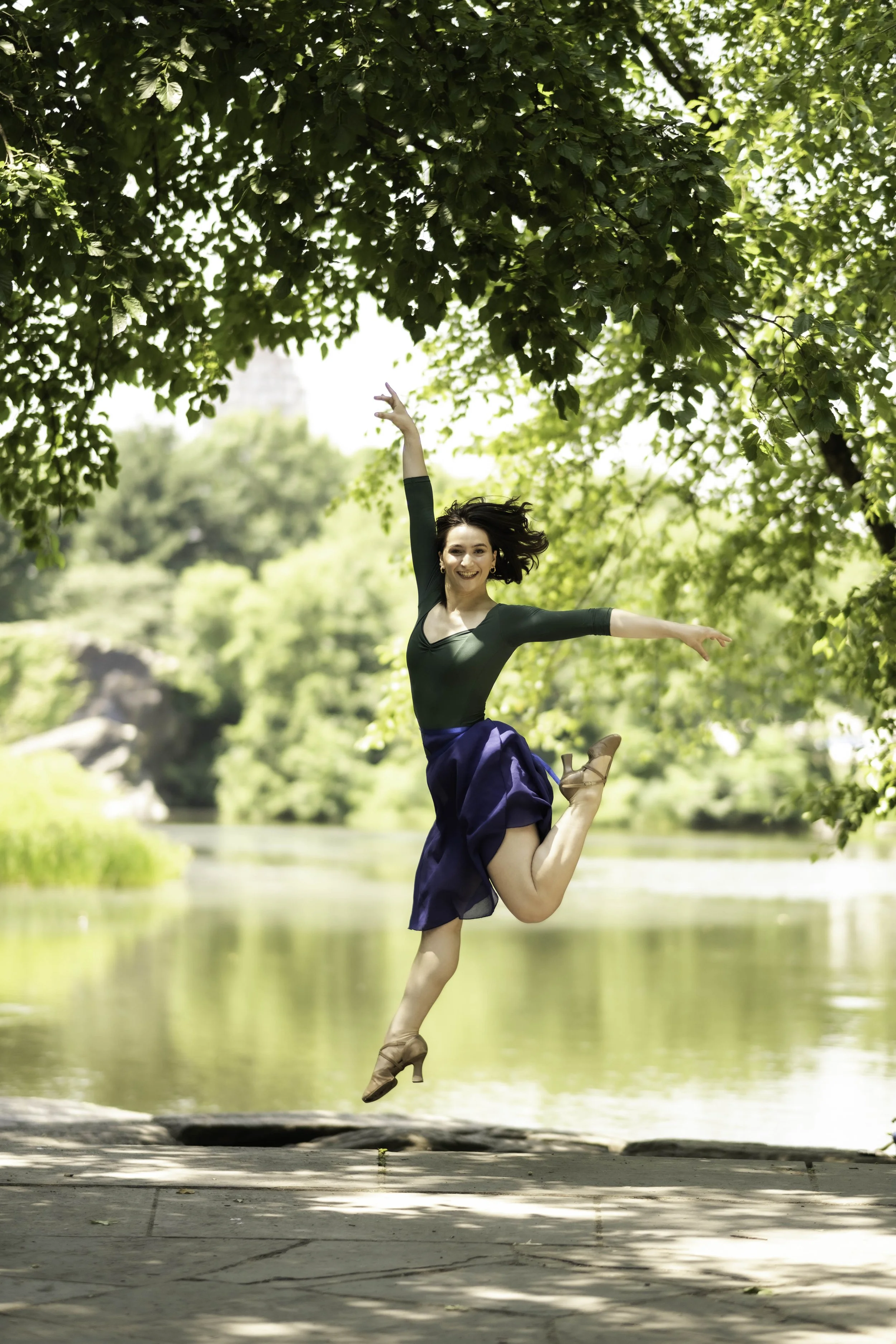 A woman ballet dancer wearing a dark green top and a blue skirt, balancing on one leg with the other bent behind her, in an outdoor setting near a river surrounded by green trees, with her arms outstretched.