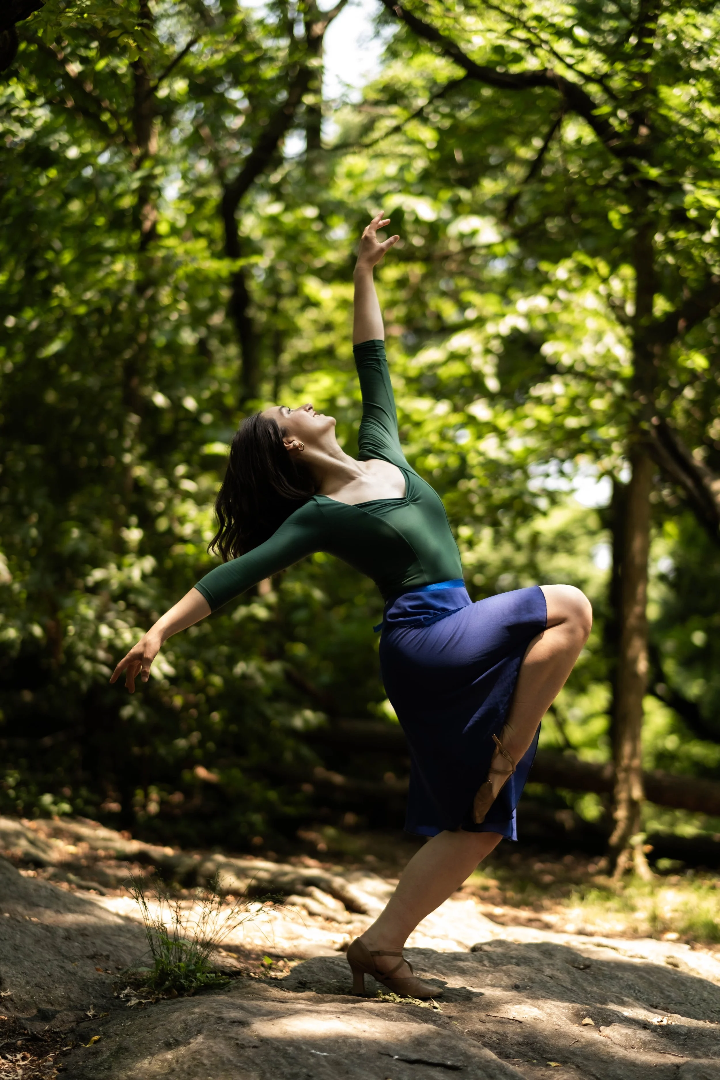 A woman practicing yoga outdoors in a forest, balancing on one leg with her arms extended, wearing a green top and blue shorts.
