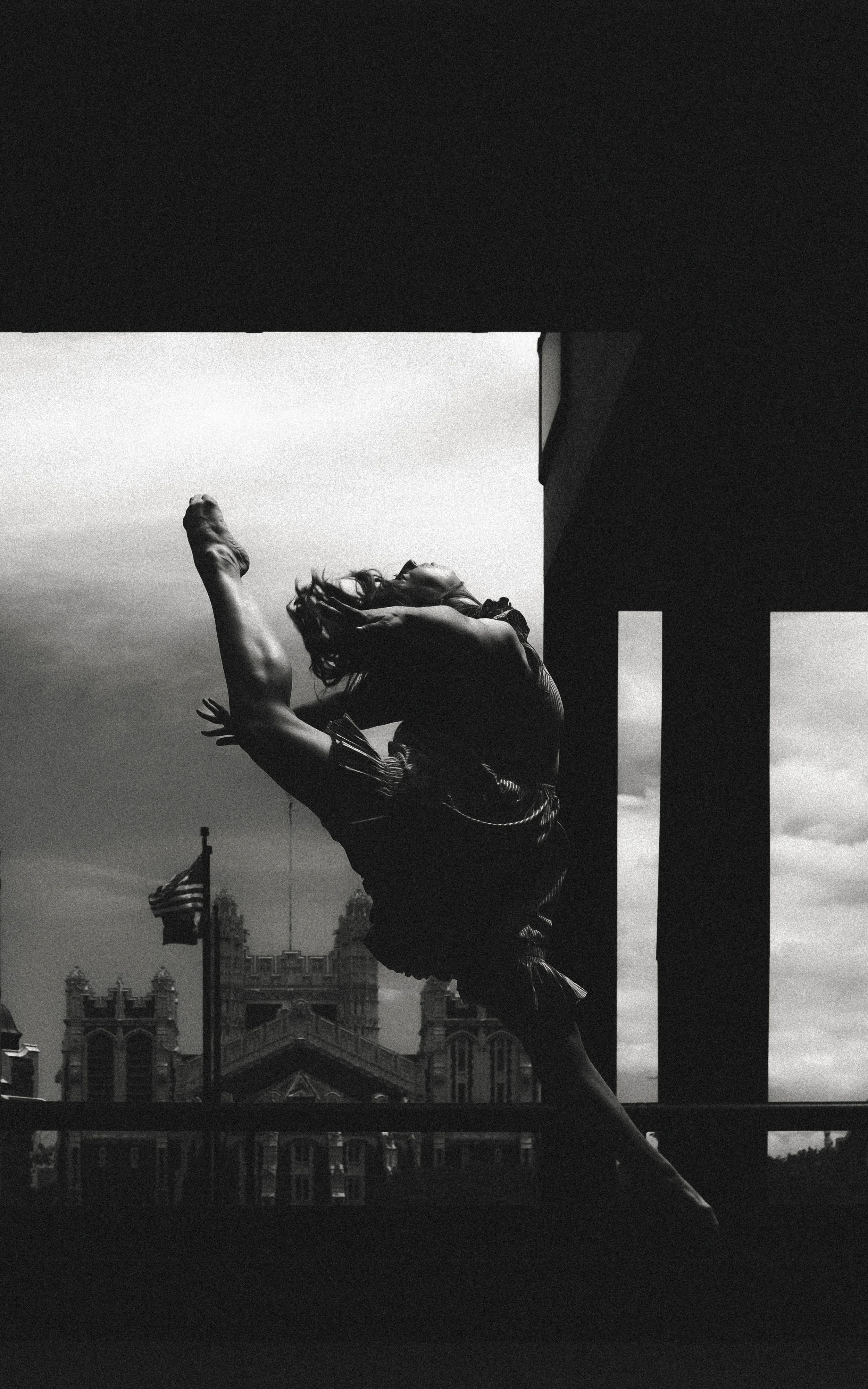 Black and white photo of a person dancing outdoors in front of a historic building and American flags, with arms raised and head tilted back.
