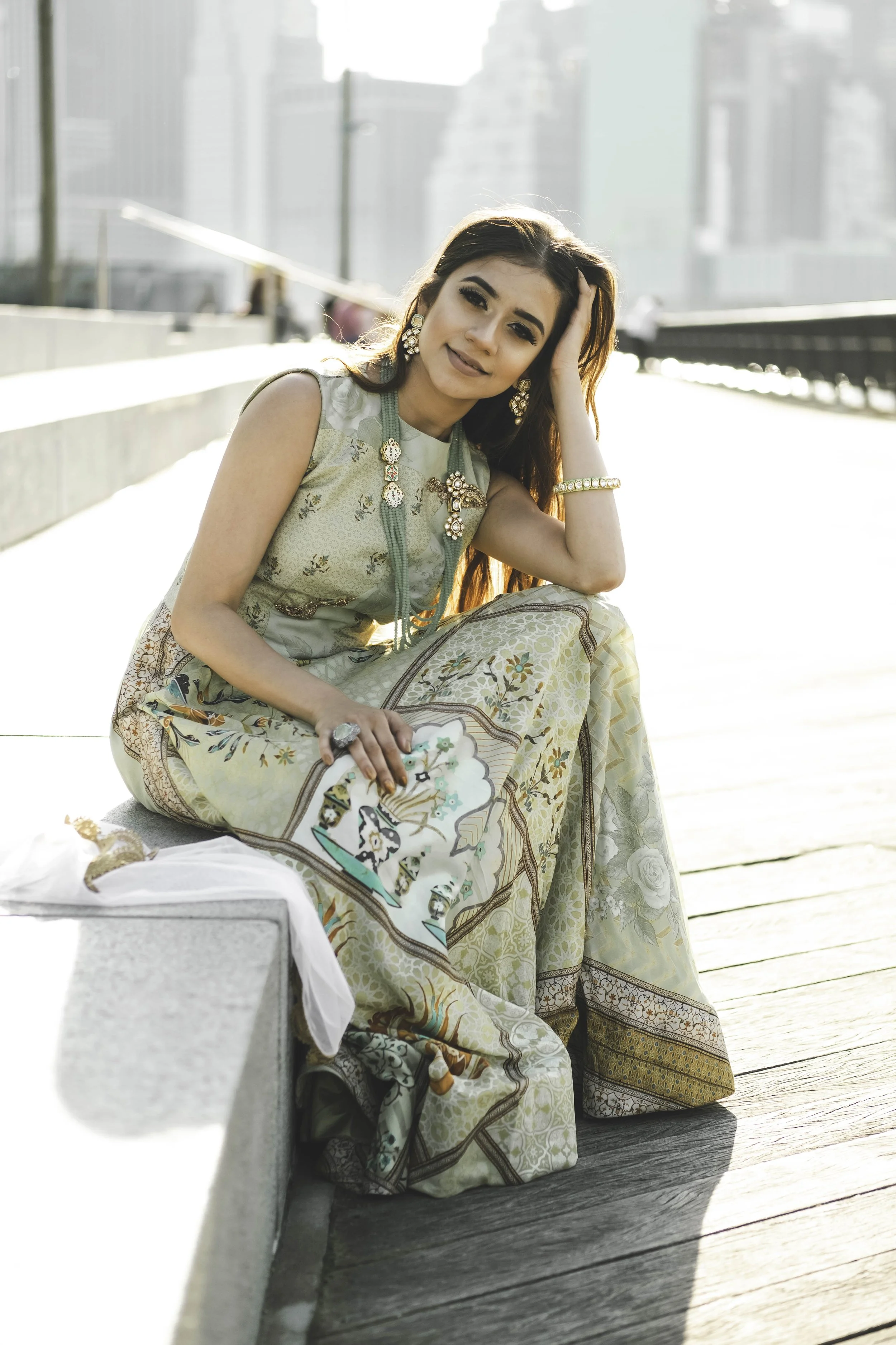 A woman with dark hair, wearing traditional dress and jewelry, is sitting outdoors on a wooden deck, with city buildings in the background, looking at the camera.
