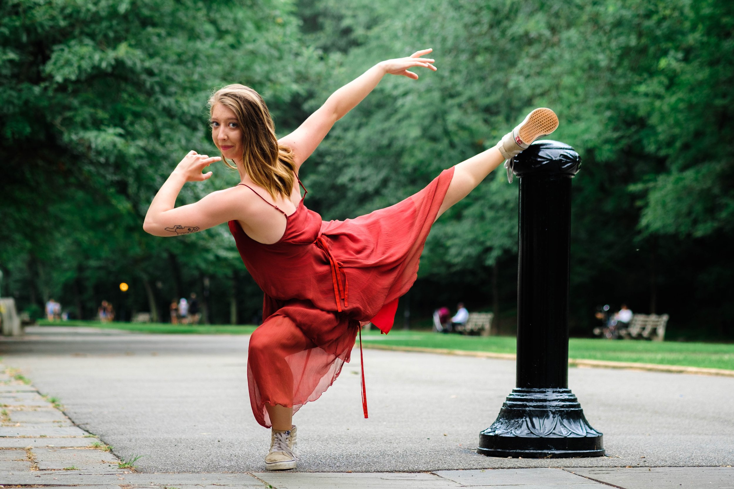 A young woman in a red dress performing a high kick, with her leg resting on a black park lamp post in a green park.