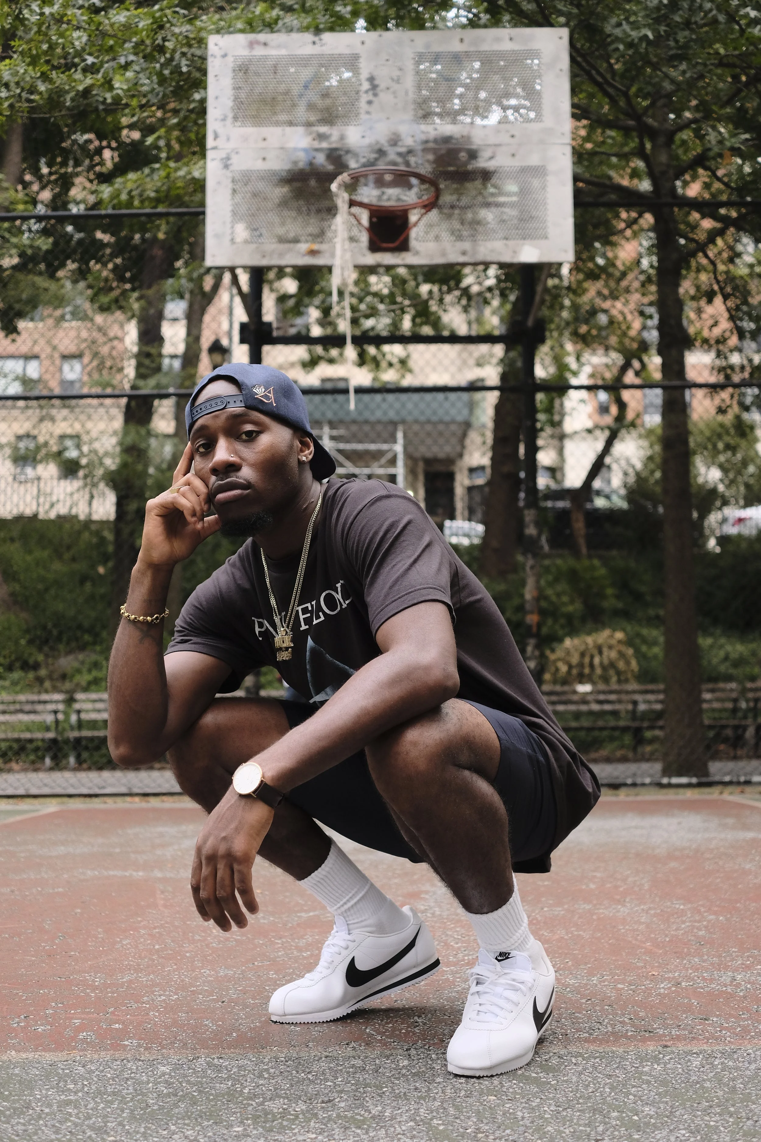 A young man squatting on an outdoor basketball court, with a basketball hoop behind him. He is wearing a dark t-shirt, black shorts, white Nike sneakers, white socks, a watch, and a gold chain. He is resting his chin on his hand and looking at the camera with a serious expression. There are trees and a building in the background.