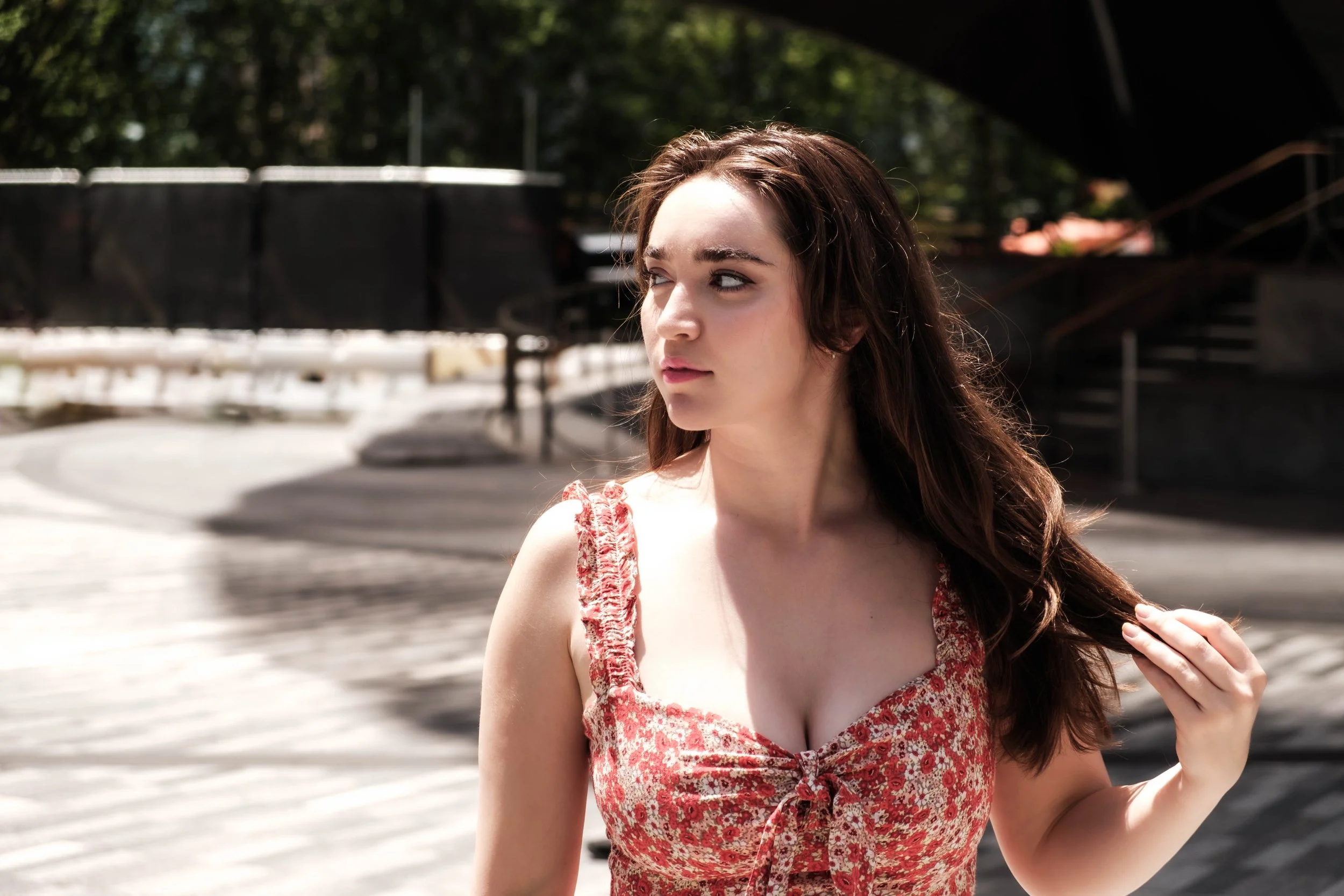 A young woman with long brown hair looking to her left, wearing a red floral dress, outdoors on a sunny day with trees and a black structure in the background.