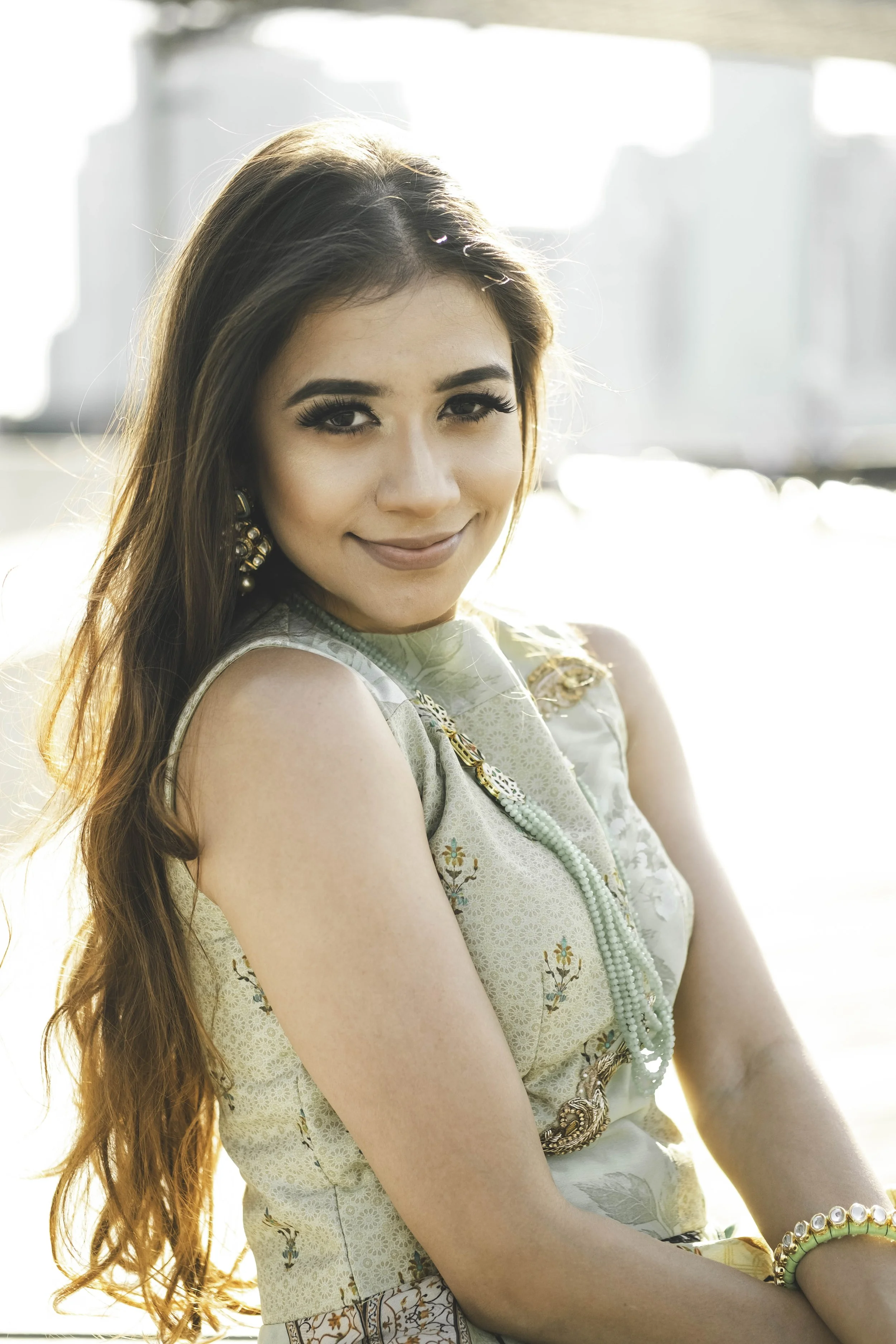 A young woman with long, wavy hair, wearing a sleeveless dress with an embroidered floral pattern, gold earrings, and layered necklaces. She is smiling softly and sitting outdoors in bright sunlight near a window or balcony with blurred cityscape background.