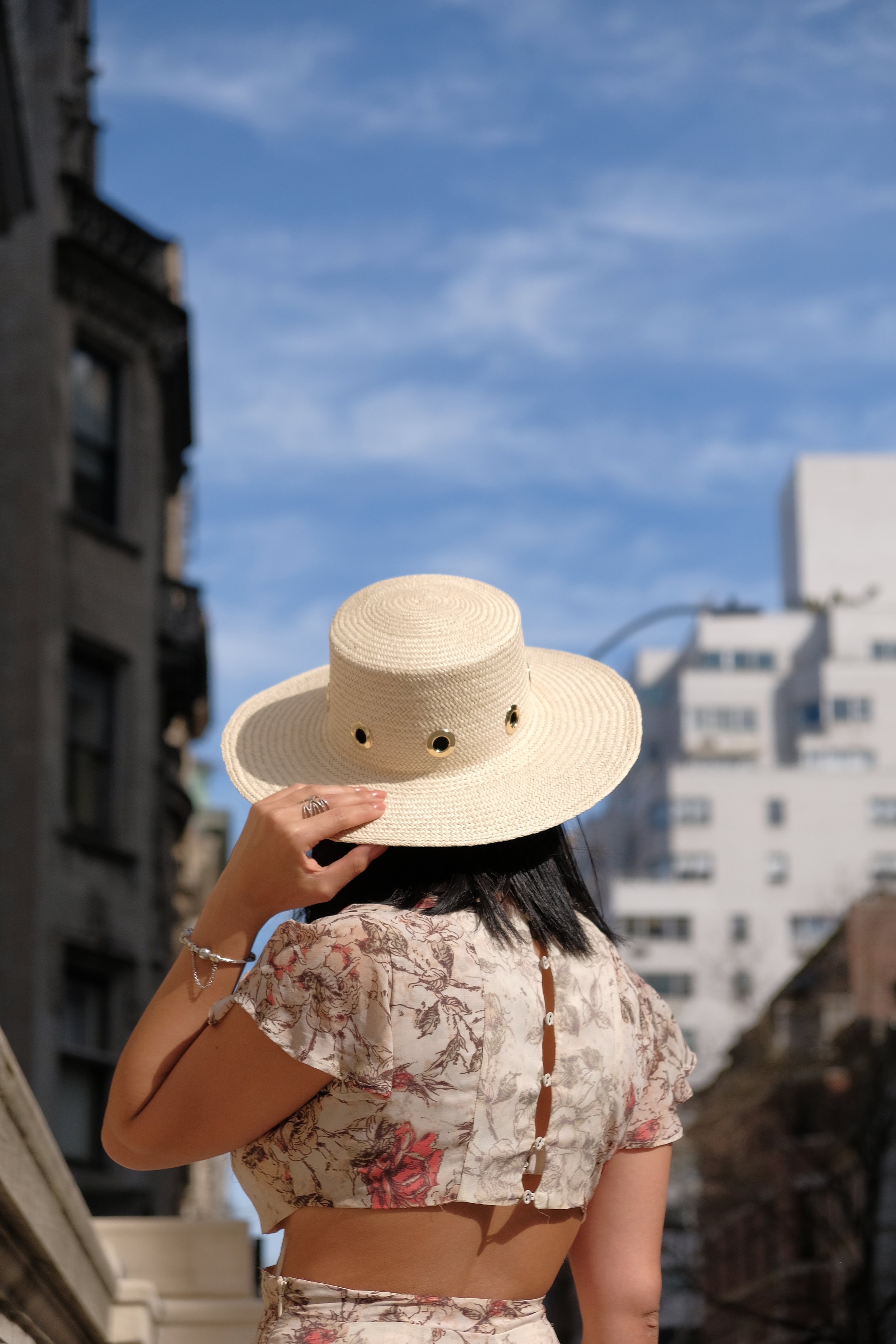 A woman holding a straw sun hat in front of her face, standing outdoors on a city street with buildings and a blue sky in the background.