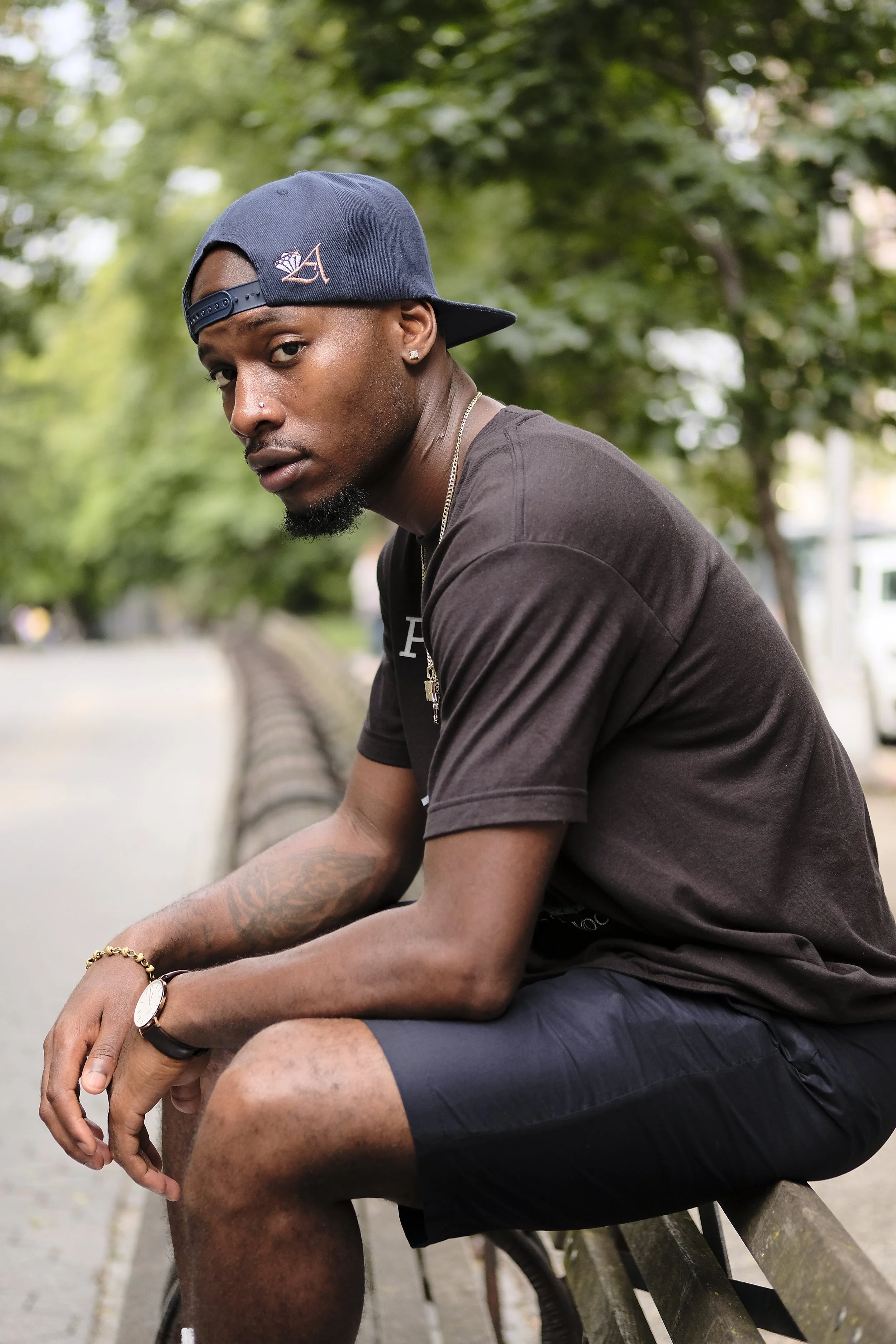 A young man sitting on a park bench, wearing a backwards baseball cap, black t-shirt, and shorts, with a tree-lined background and a blurred cityscape in the distance.