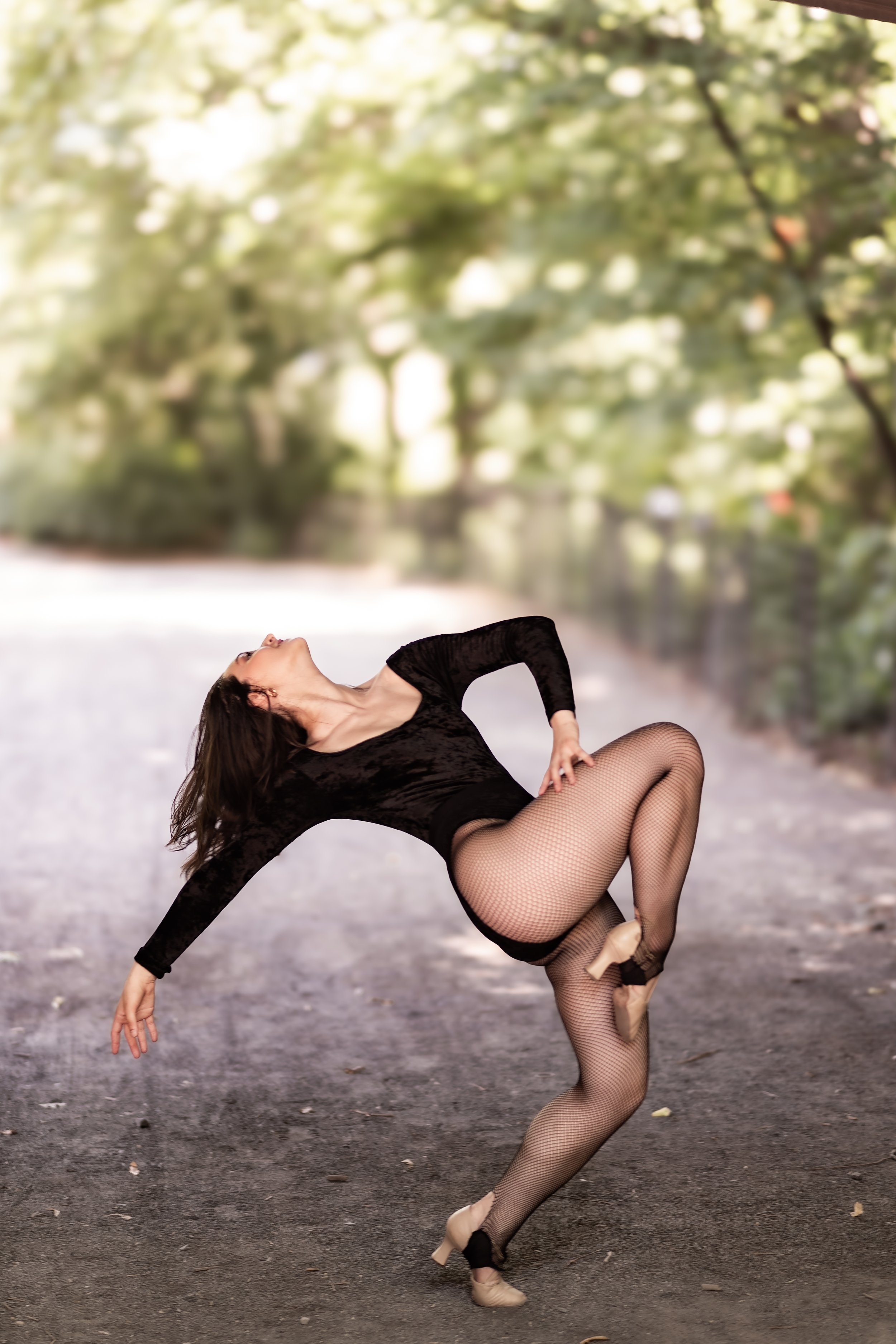 A woman in black dance attire and fishnet tights performing a dance pose outdoors on a dirt path with blurred green trees in the background.