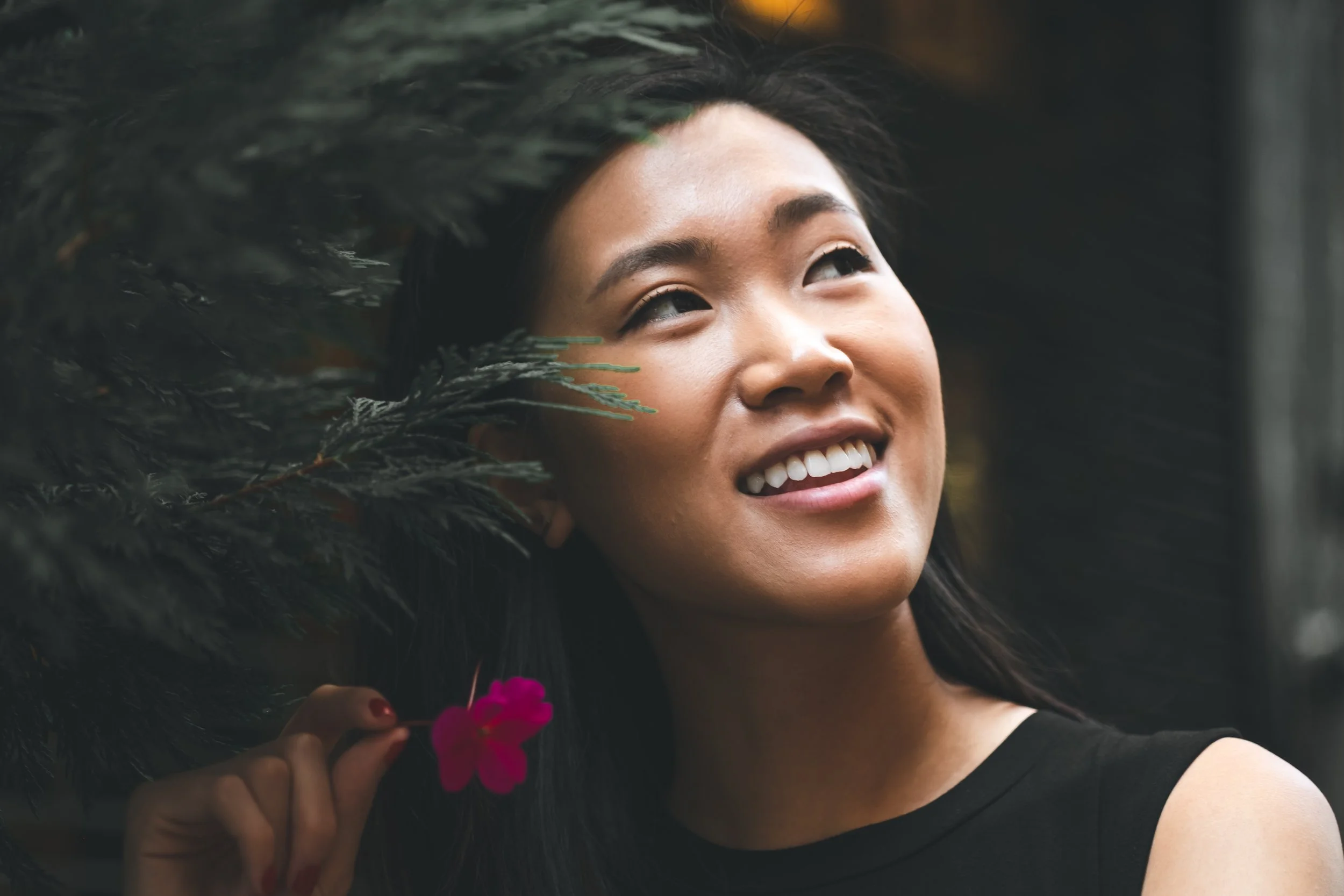 A close-up of a smiling woman with dark hair, surrounded by greenery, holding a pink flower.