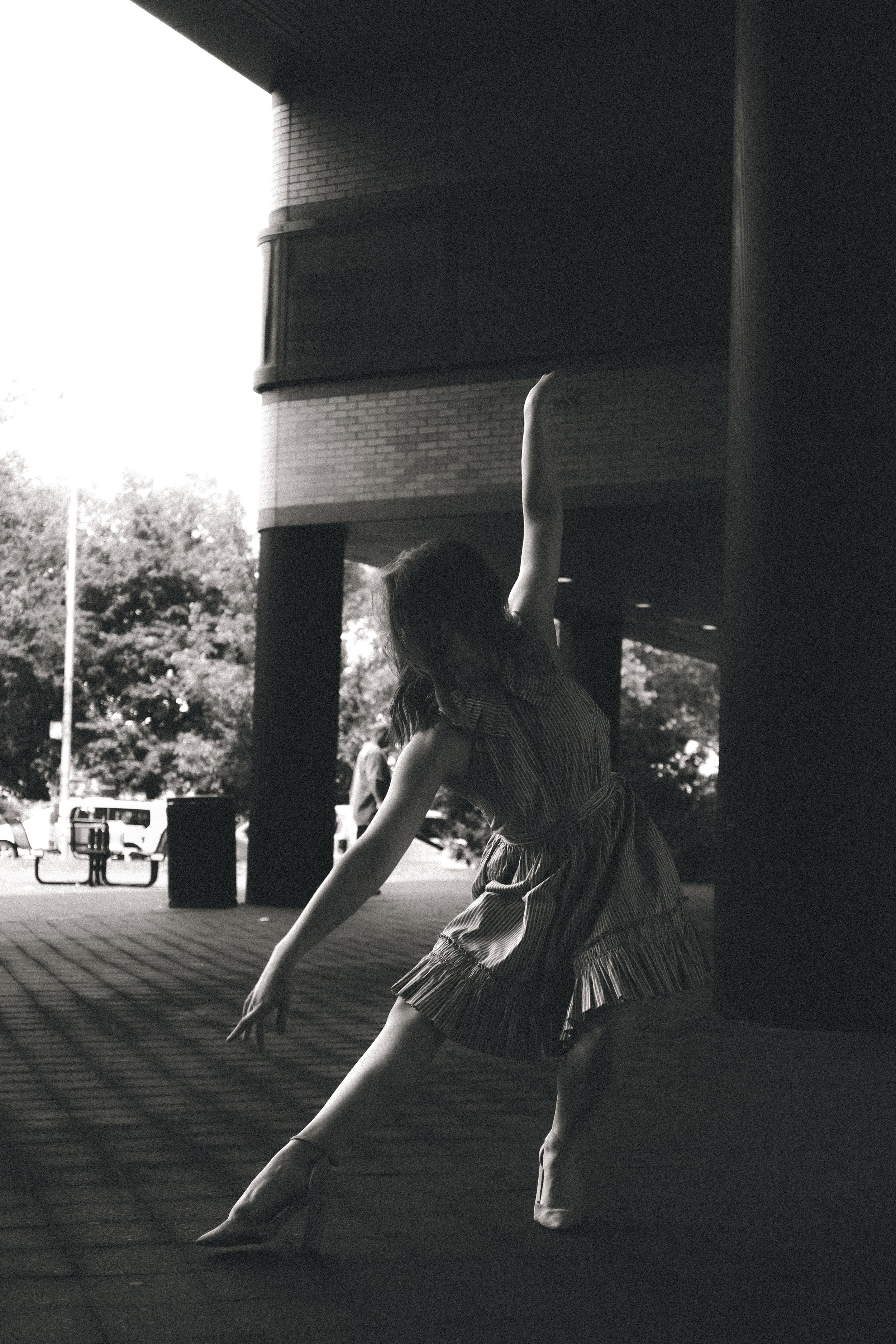 A woman with short hair wearing a striped dress and pointed shoes is dancing or performing ballet outdoors. She is reaching her right arm upward and leaning forward, with her left arm extended downward. The background shows trees, a brick building, a