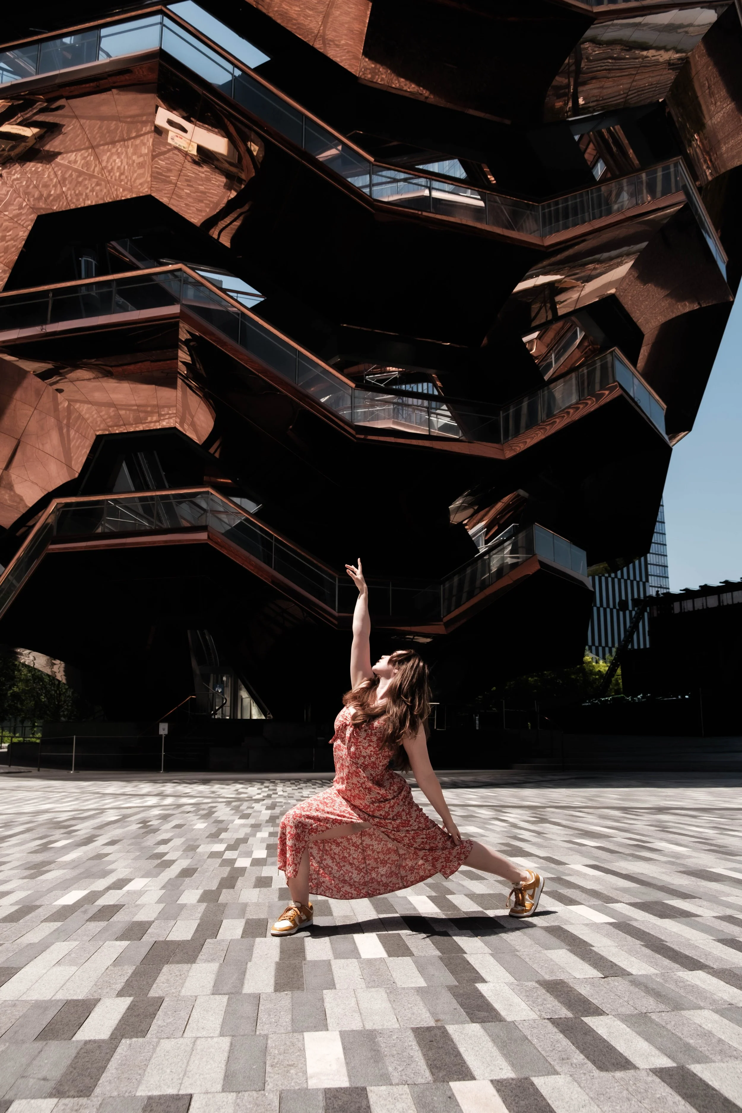 A woman in a floral dress and sneakers poses in a dance pose on a patterned tiled floor in front of an modern, architectural building with multiple levels, glass windows, and dark exterior.