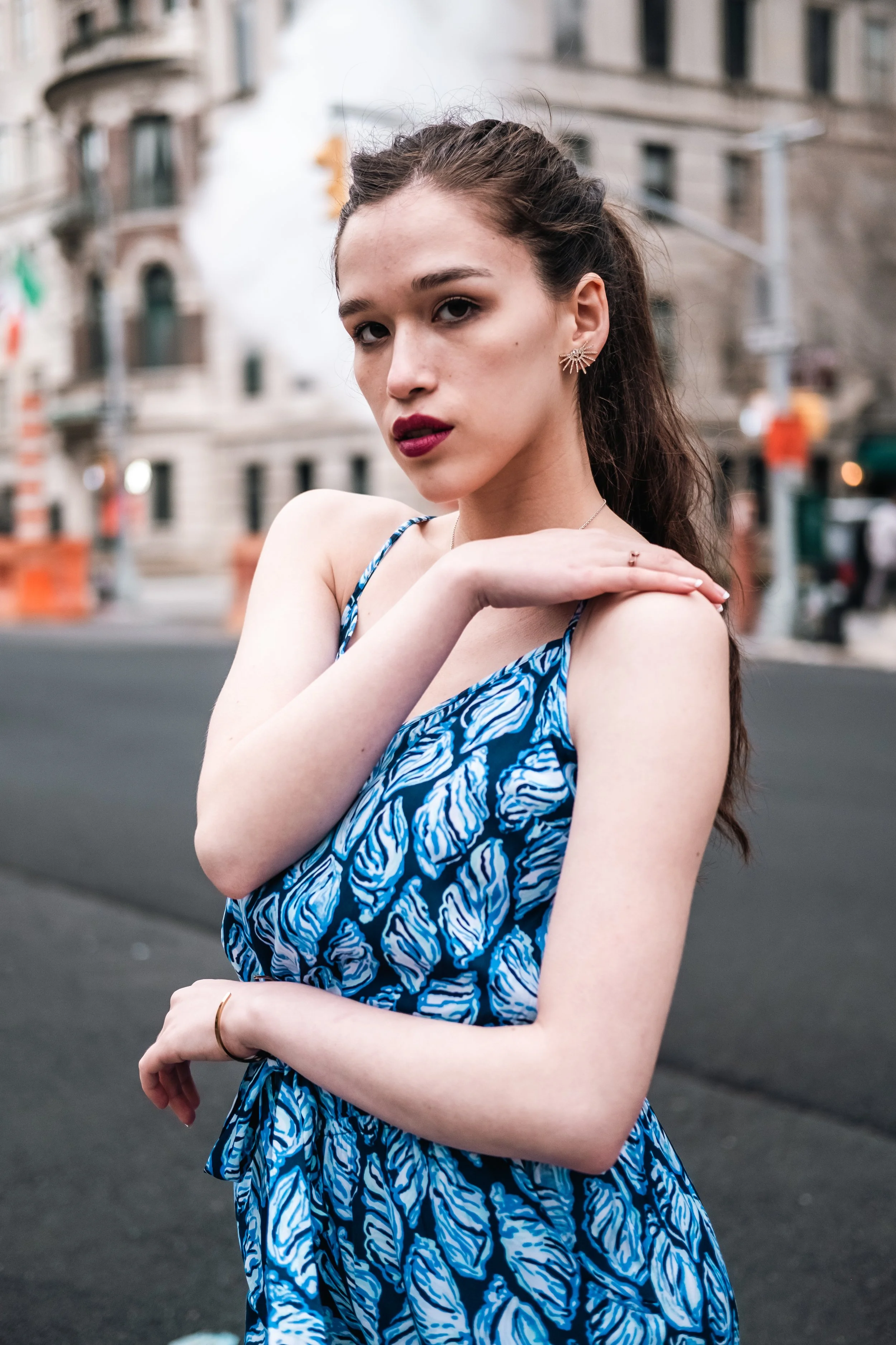 A young woman with dark hair styled in a high half-pony, wearing a blue patterned dress and statement earrings, posing outdoors on a city street during daytime.