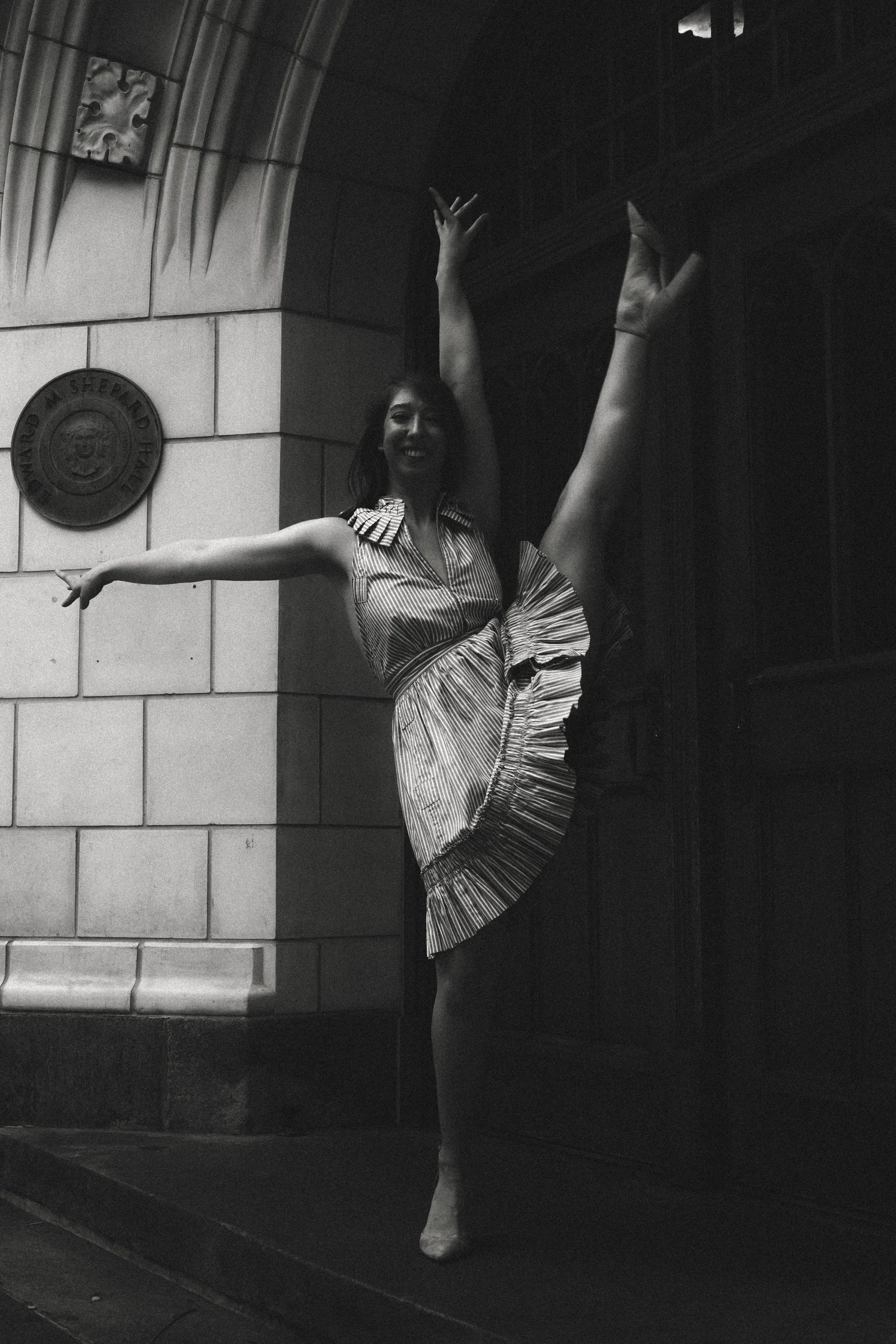 A woman in a striped dress performing a ballet pose with one leg extended vertically, against a background of a brick wall and decorative stonework.