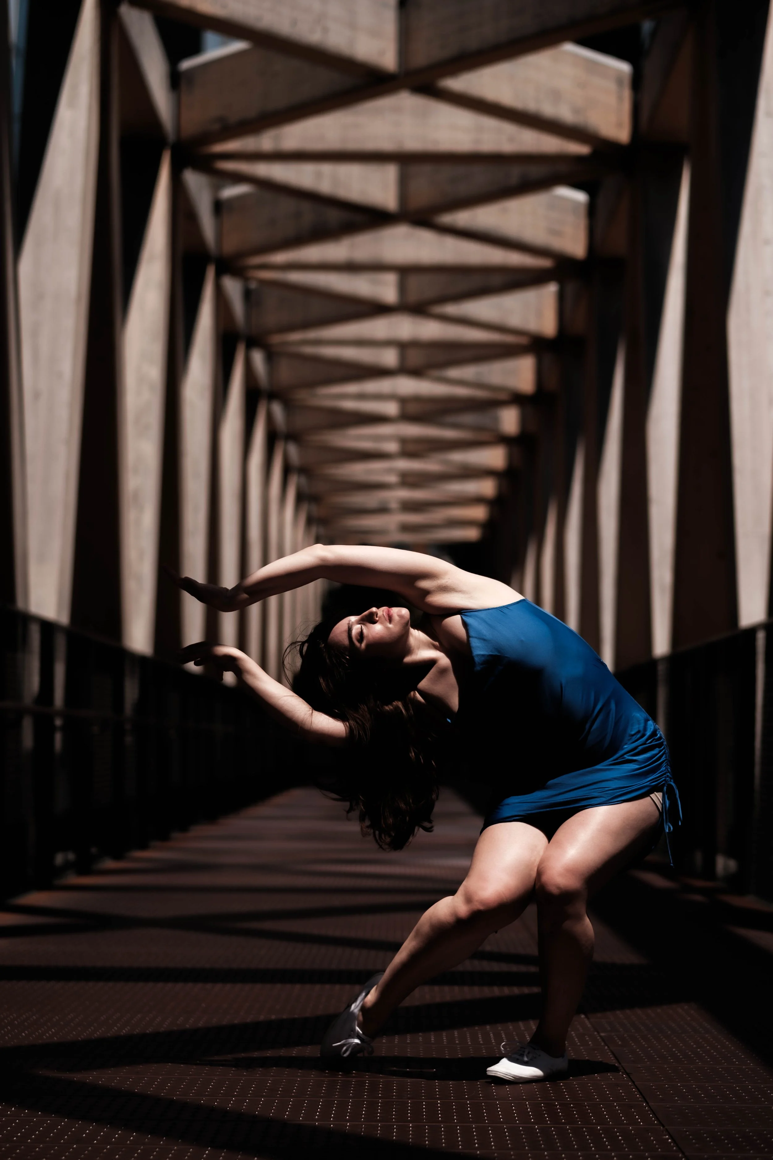A woman in a blue athletic dress performing a yoga pose in a dimly lit industrial-style hallway with geometric wooden ceiling beams.