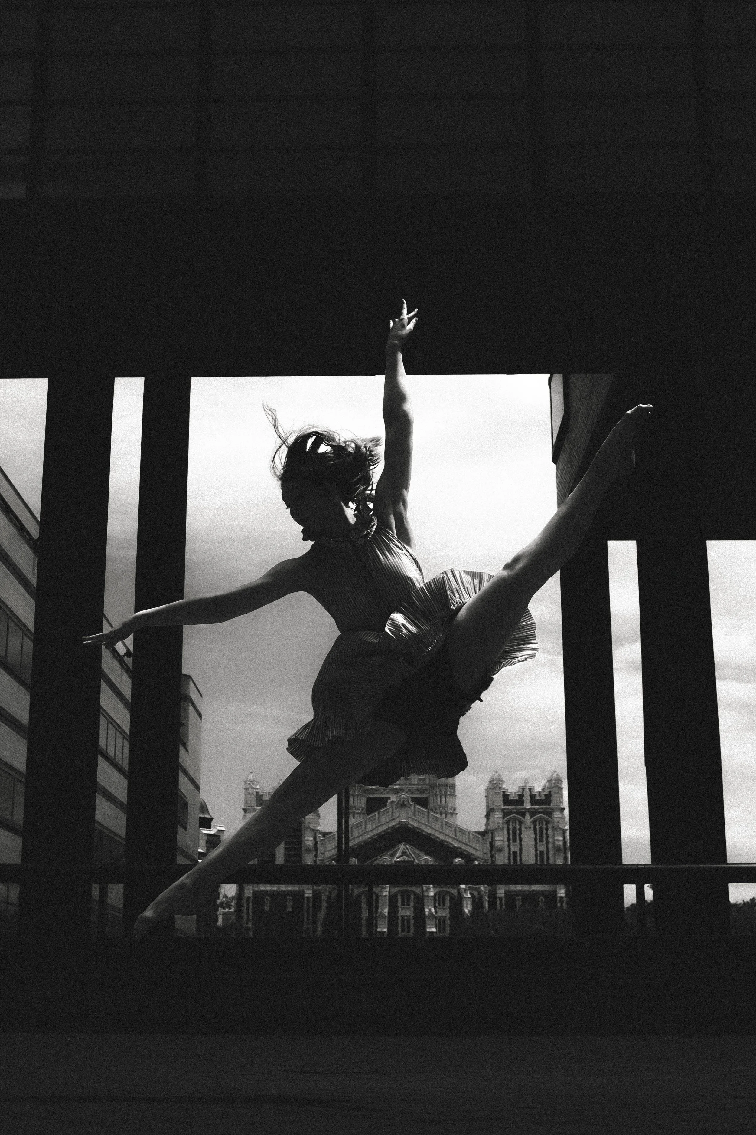 A woman is leaping through the air with her arms and legs extended, dancing or performing an artistic jump, with a cityscape and historic buildings in the background. The photograph is in black and white.