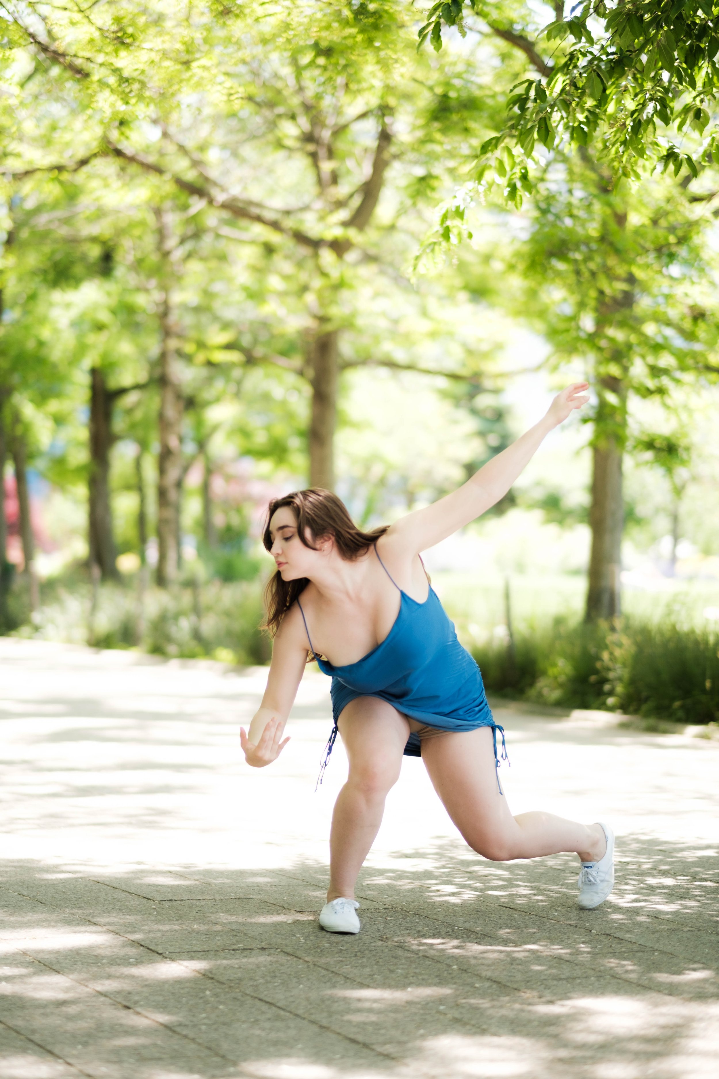 A young woman in a blue dress and white sneakers practicing yoga outdoors on a sunny day, surrounded by green trees.