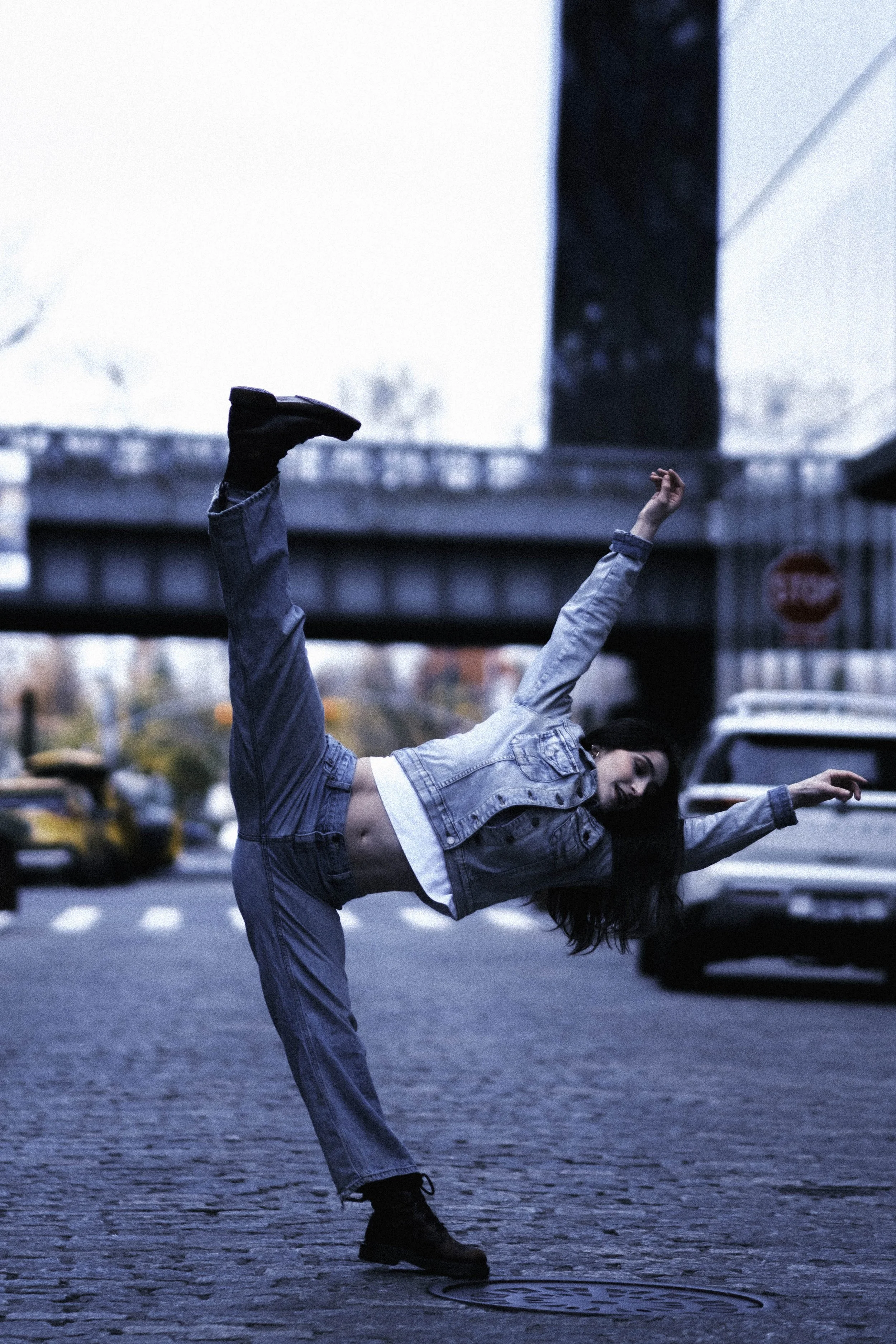 A woman wearing a denim jacket and jeans is performing a dance or pose on a city street, with her arms extended and her left leg lifted high. The background shows a traffic scene with cars and a large building, suggesting an urban environment.