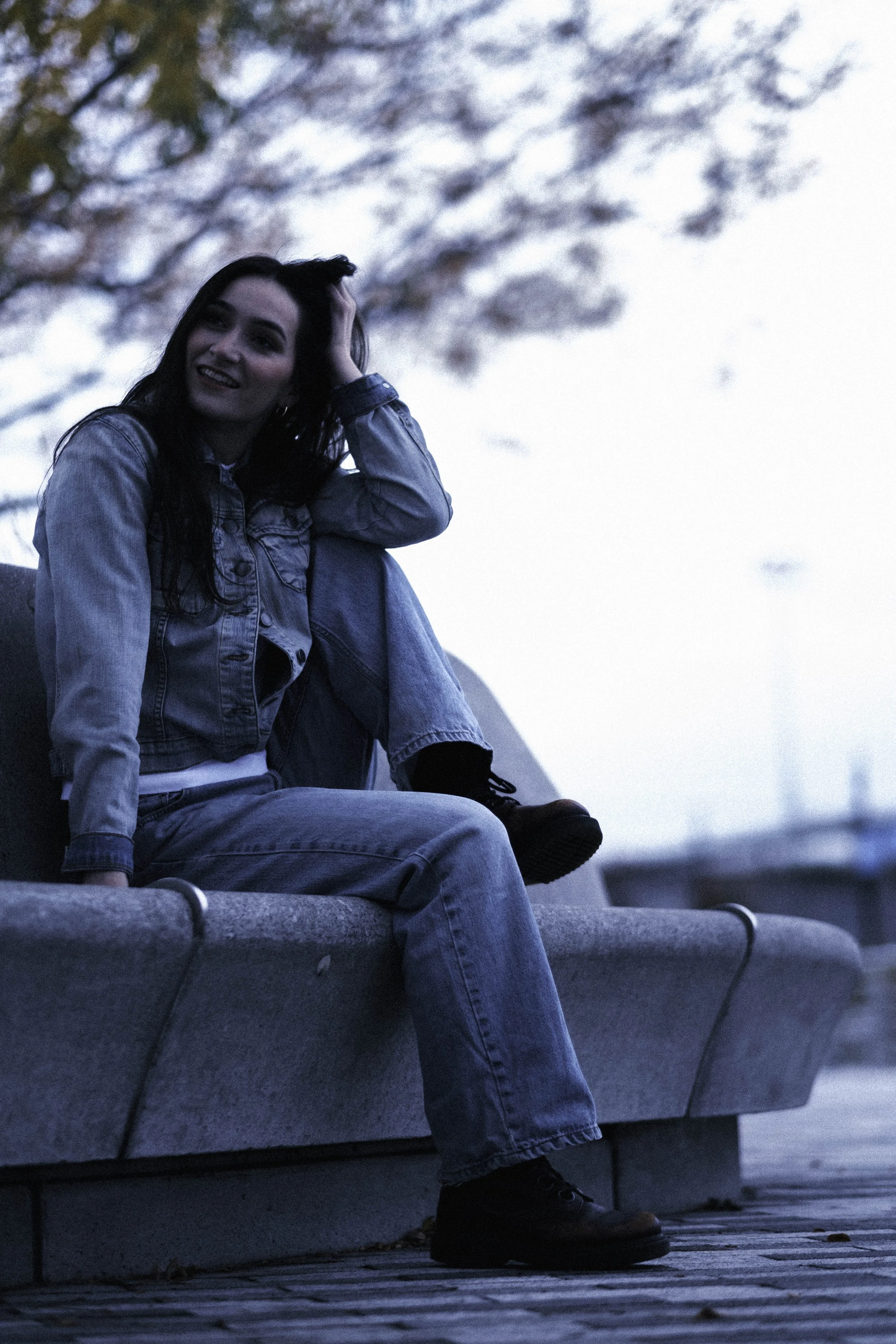 A young woman with dark hair sitting on a concrete bench outdoors, wearing a denim jacket and jeans, smiling and touching her head, with trees and sky in the background.