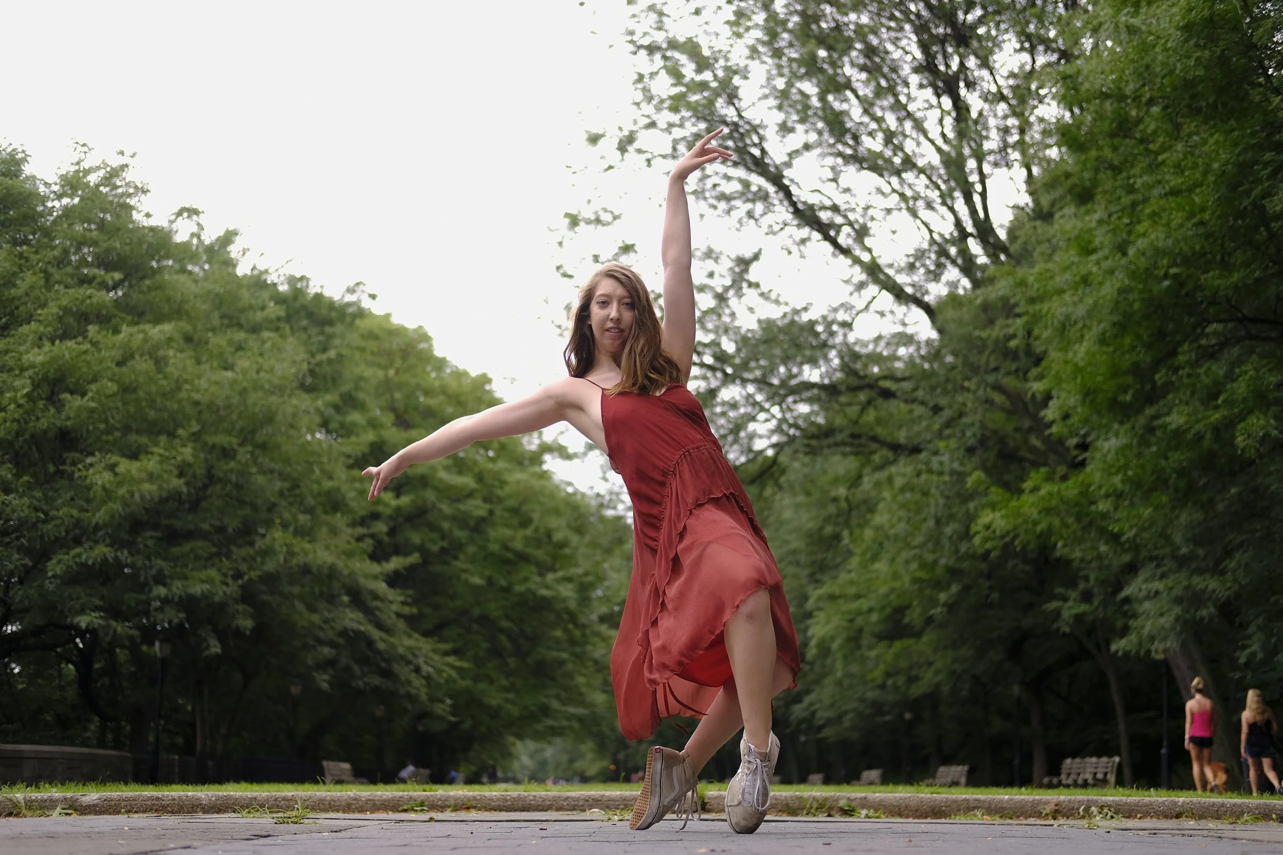 A young woman in a red dress dancing outdoors on a paved path in a park, with green trees in the background.