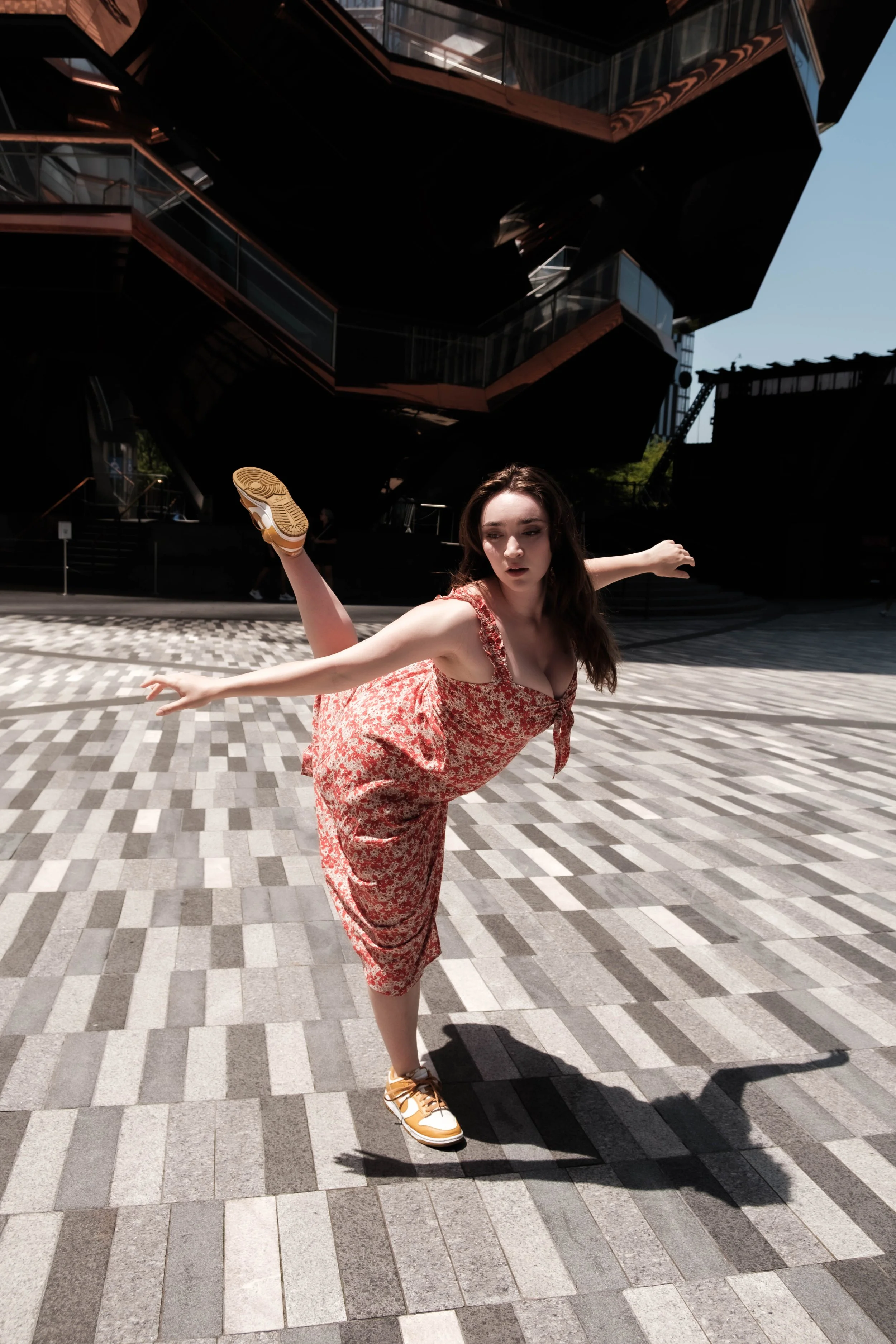 A woman in a floral dress is balancing on one leg with her arms extended, standing on a patterned outdoor surface in front of a modern, dark building with multiple levels and glass balconies.