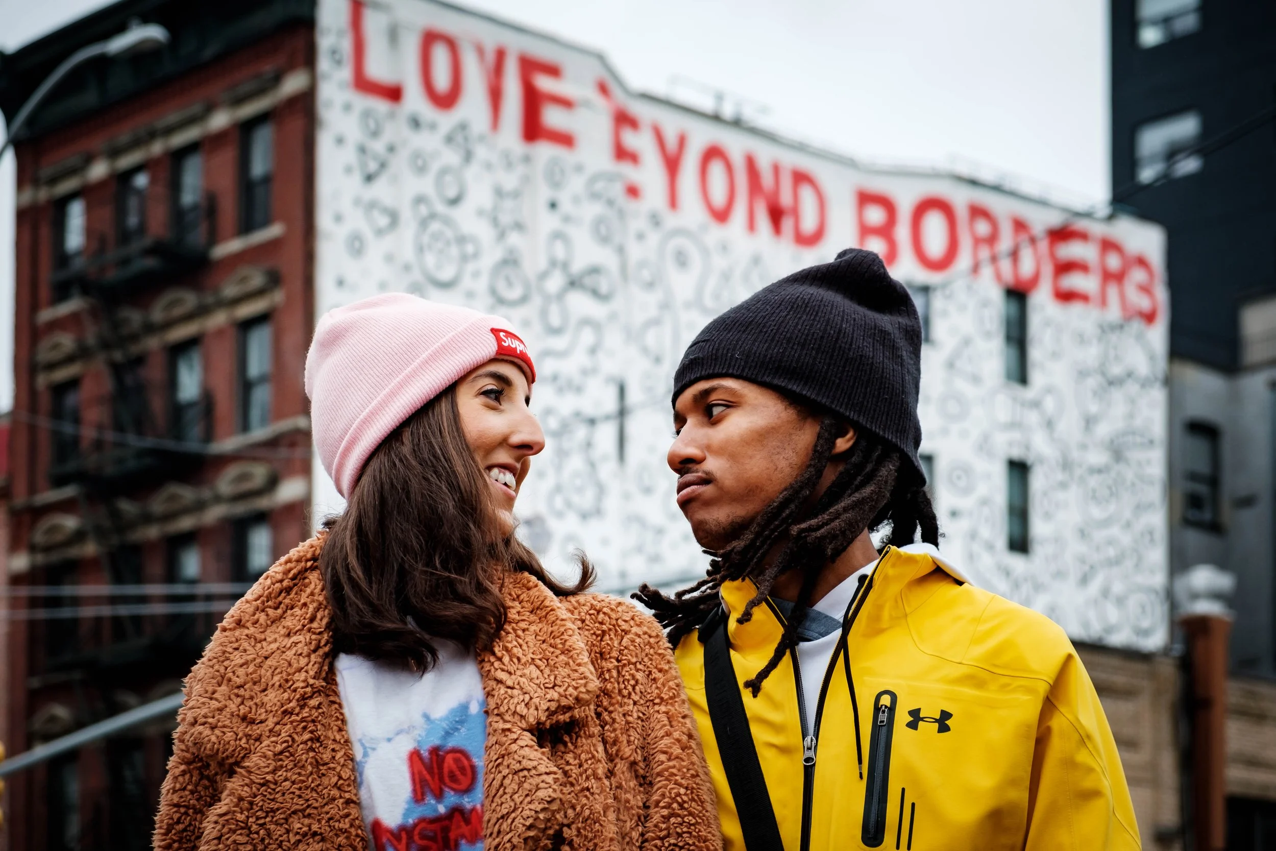 A young couple looking at each other, standing outdoors in front of a mural that reads 'LOVE YON AND BORDER' and features abstract shapes. The woman wears a pink beanie and a cozy brown jacket, while the man wears a black beanie and a yellow jacket.