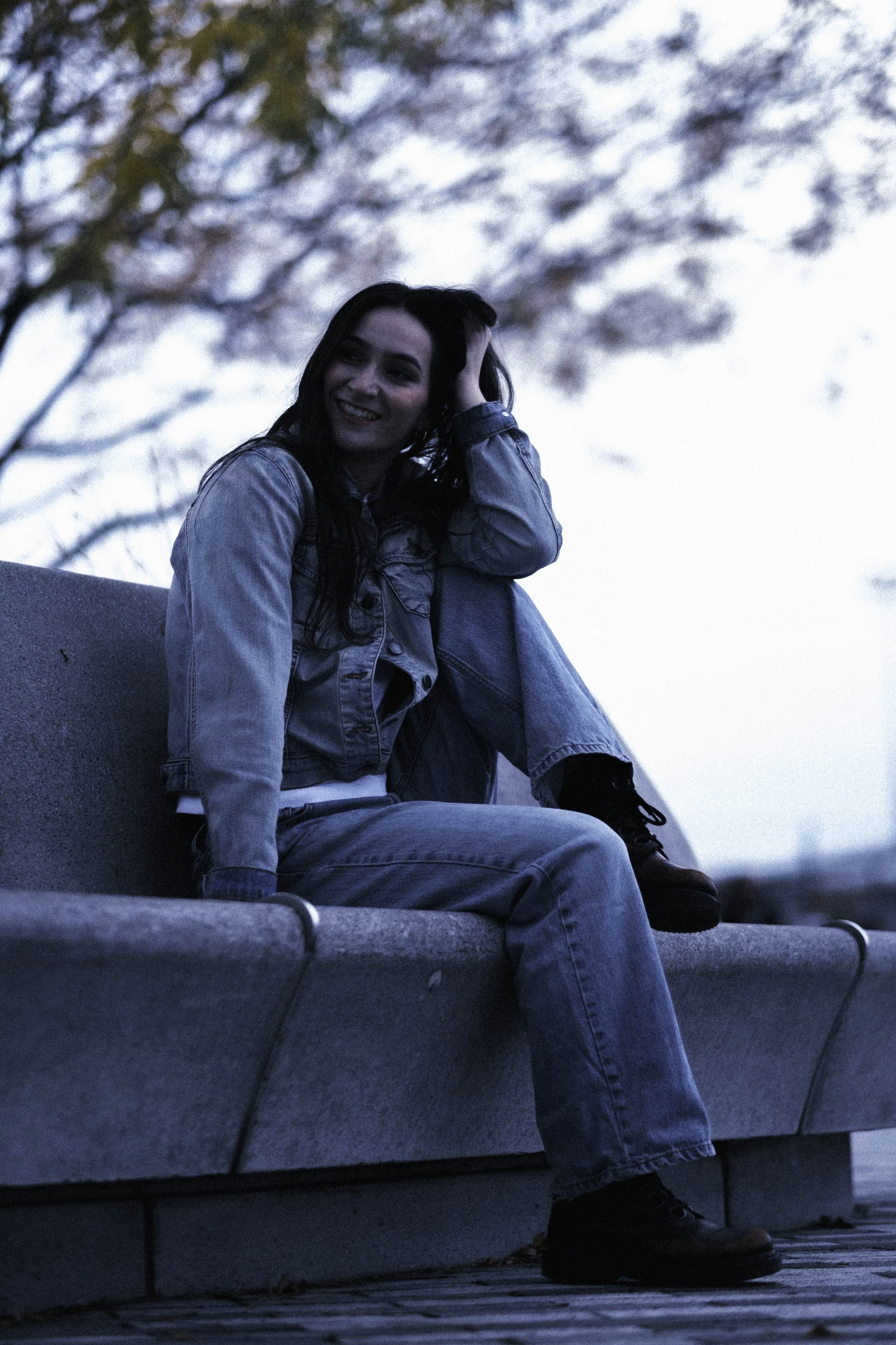 A young woman in a denim jacket and jeans sitting on a park bench during dusk, with her left hand on her head, smiling and looking to her left, with blurred trees in the background.