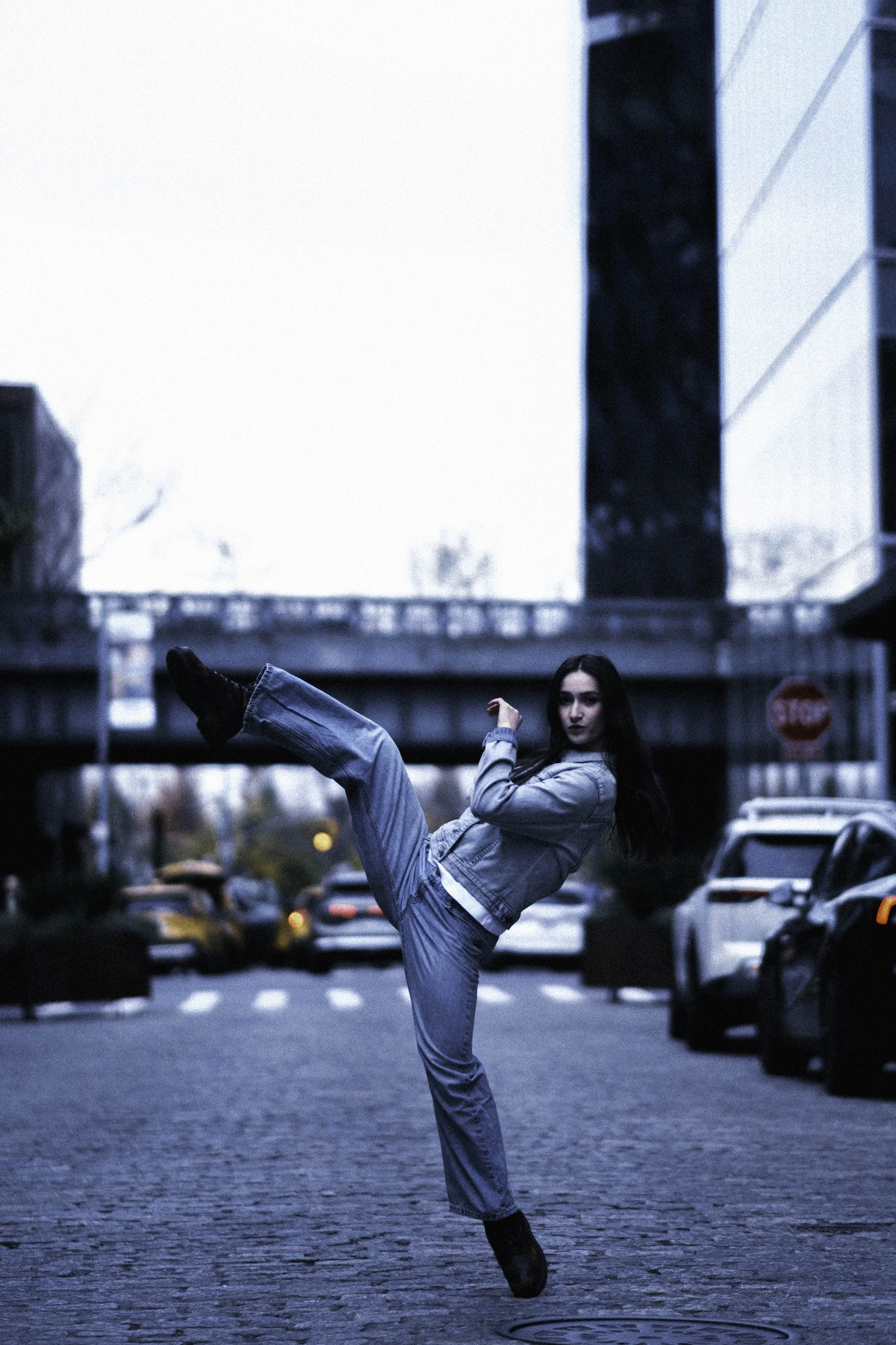 A woman dancing ballet on a city street during dusk, dressed in casual jeans and a denim jacket, with her leg extended in a high kick and dark hair.