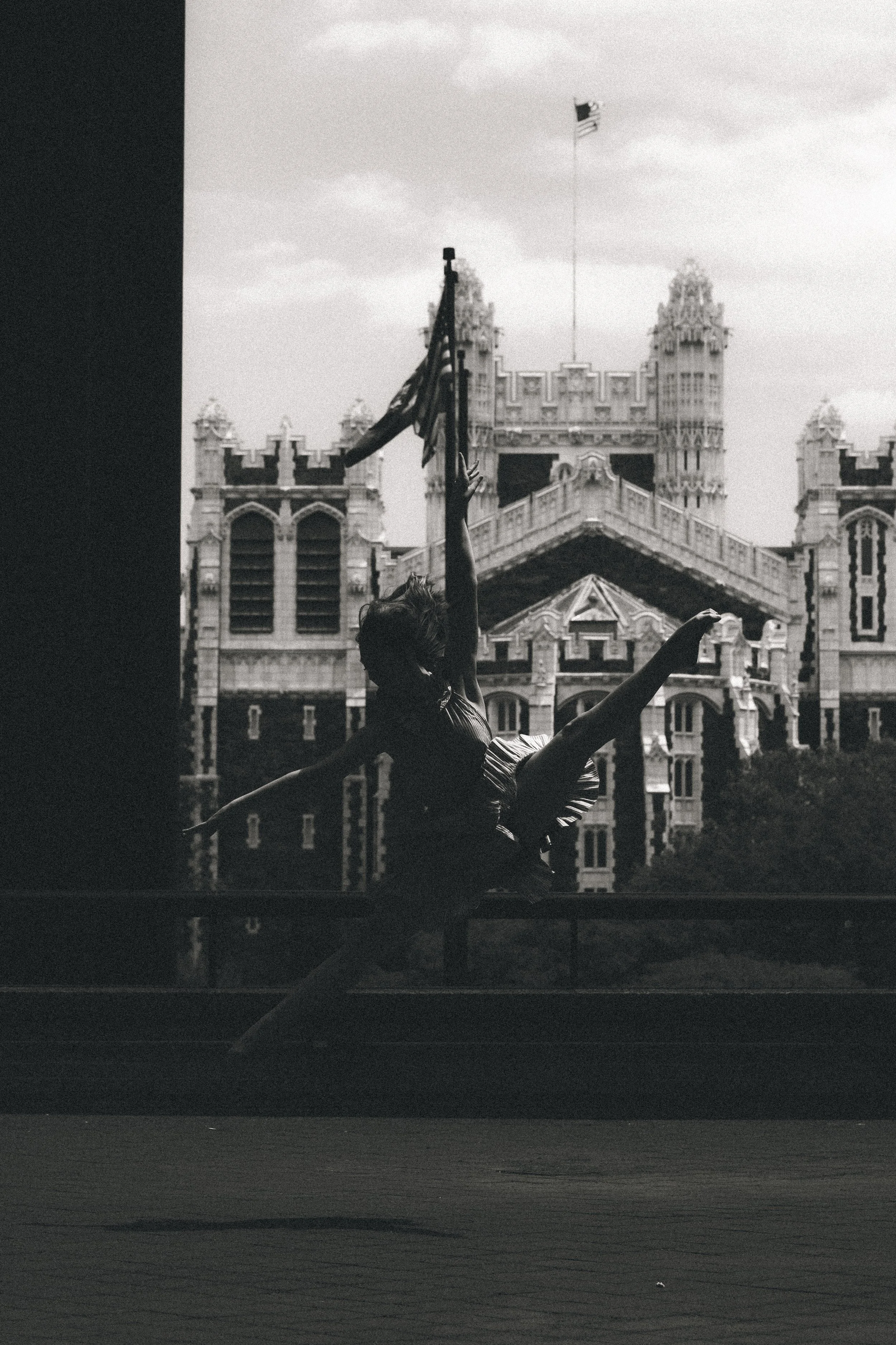 A ballerina performs in front of a historic building with a clock tower and American flags under a cloudy sky.