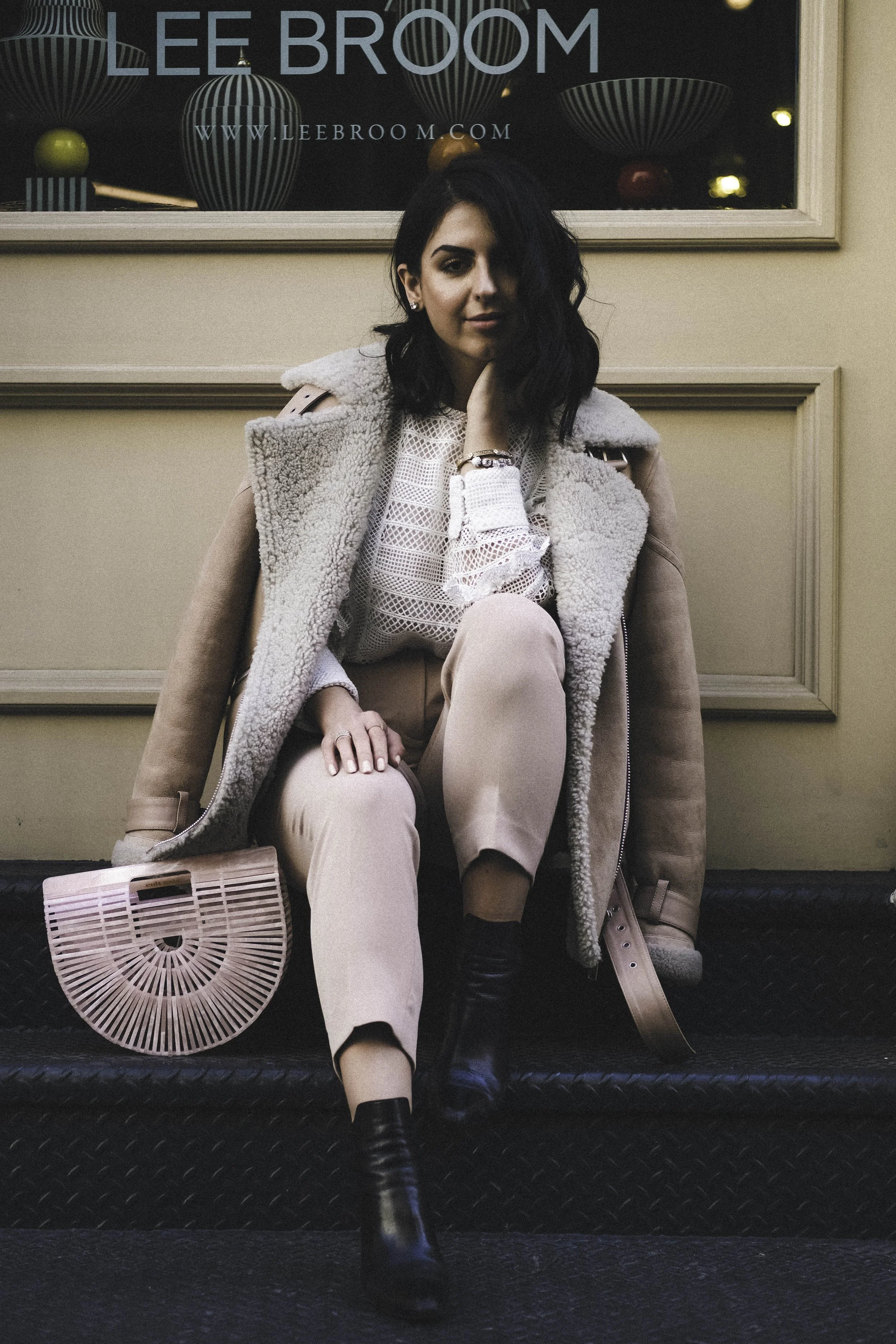 A woman with dark hair and light skin sitting on the floor outside a store with a sign for 'Lee Broom.' She wears a beige coat, a white lace top, beige pants, and black boots. She has a bracelet and earrings, and she holds a distinctive pink handbag.