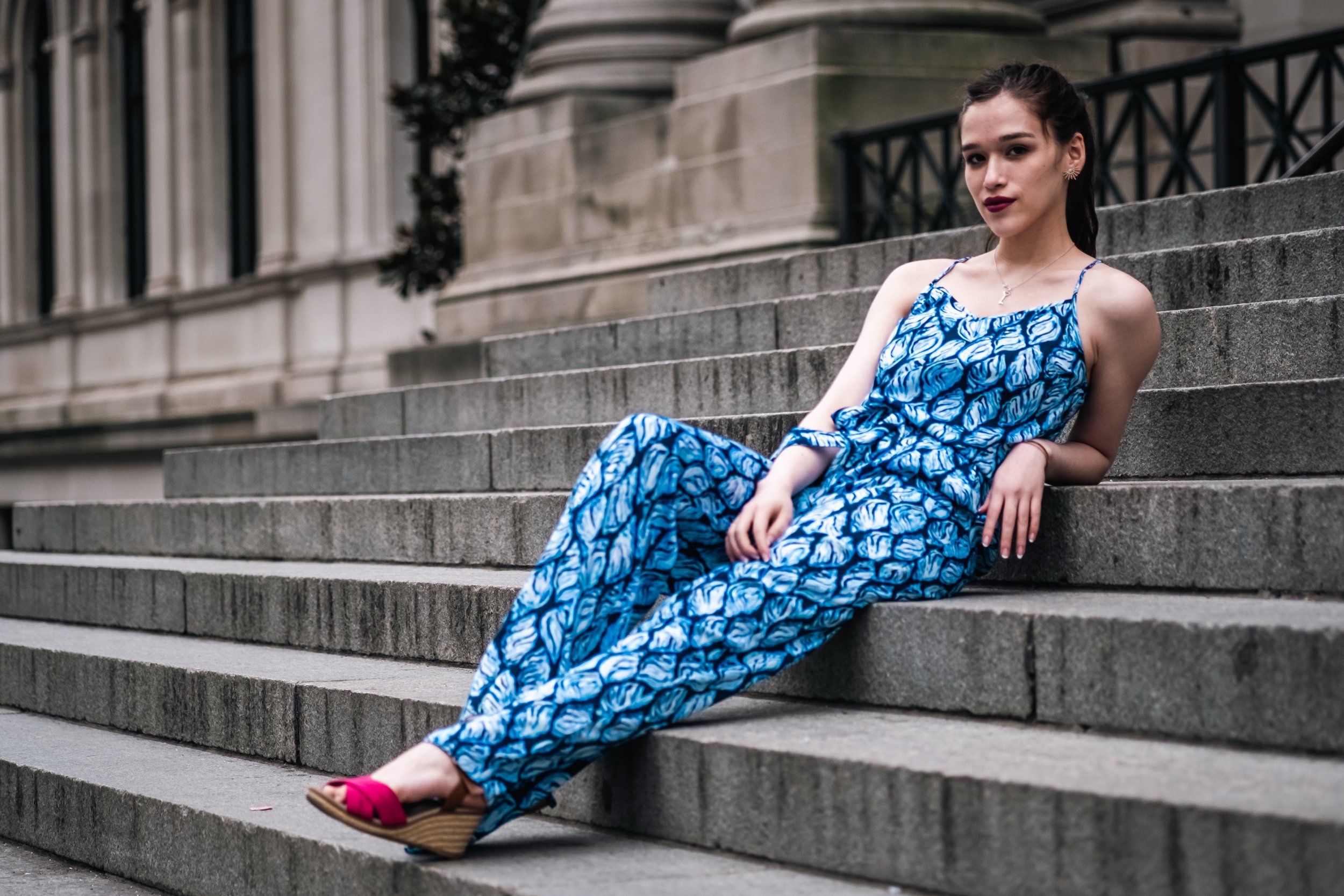 A young woman with dark hair and dark lipstick is sitting on stone steps in front of a historic building. She is wearing a blue patterned dress with spaghetti straps and pink sandals.