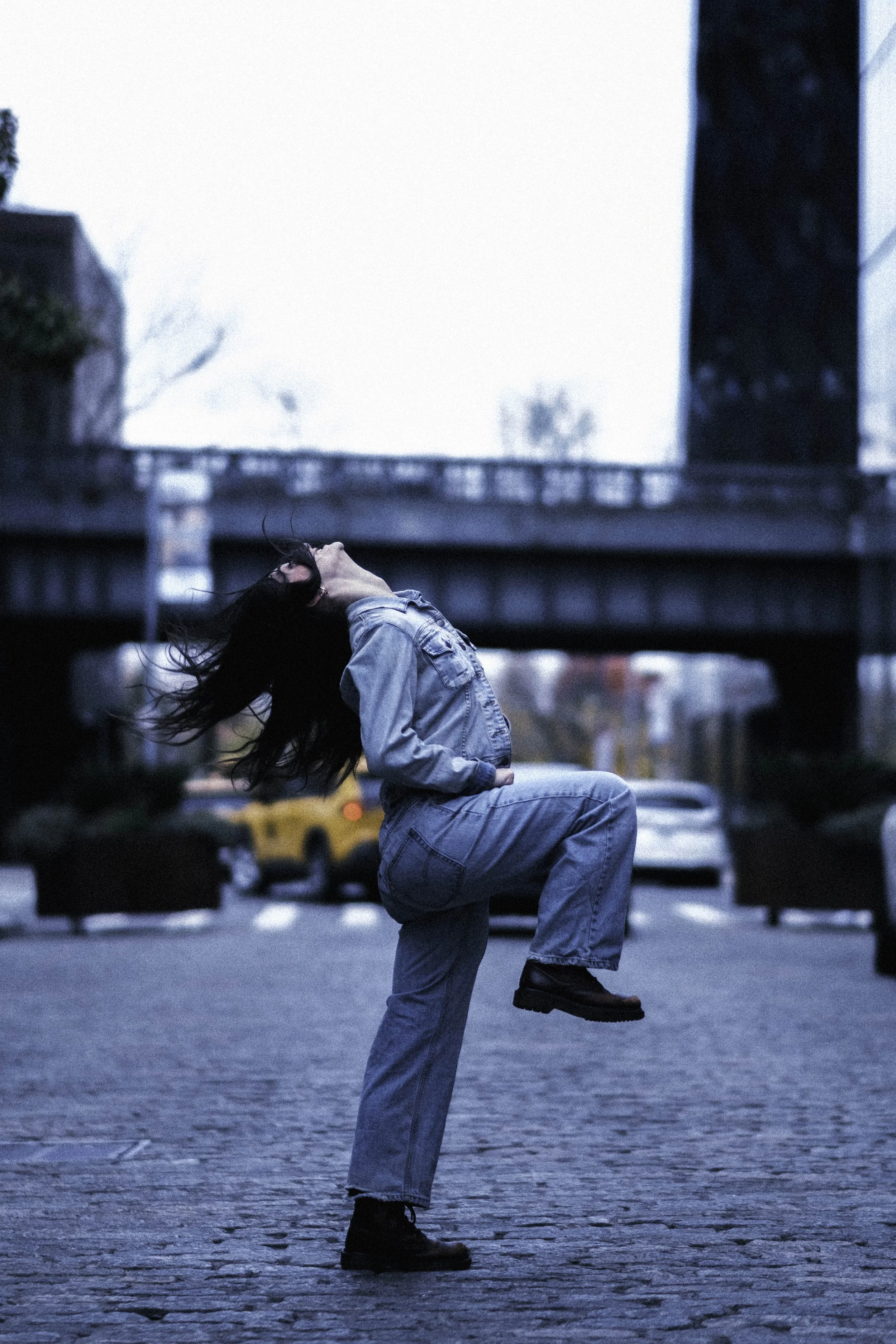 A woman with long black hair, wearing a denim jacket, jeans, and black boots, is squatting on a cobblestone street while holding her head with one hand and lifting her leg with the other. Urban background with blurred cars and a bridge.