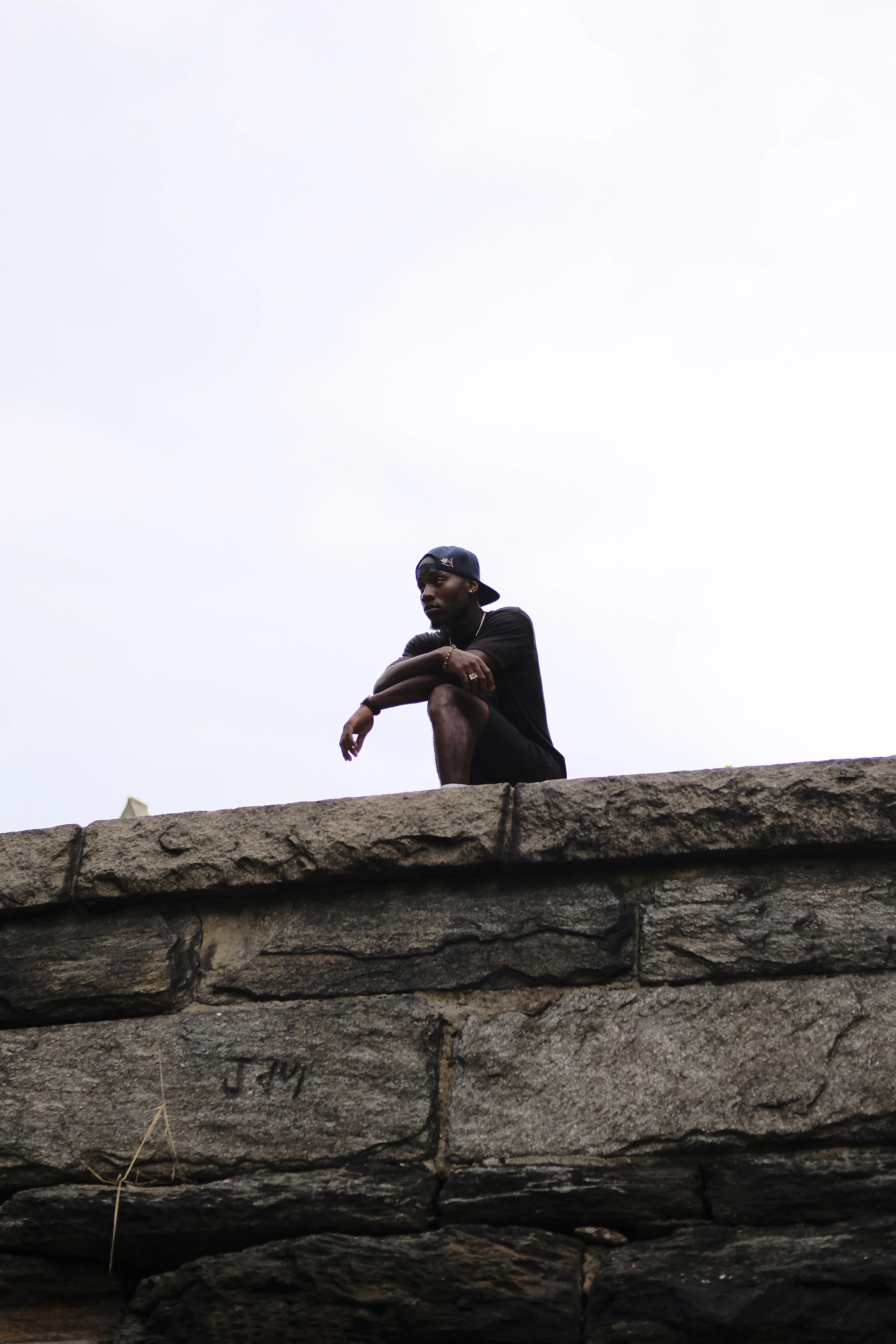 A man wearing a black t-shirt, denim cap backwards, and jewelry, crouching on a stone ledge with a cloudy sky in the background.