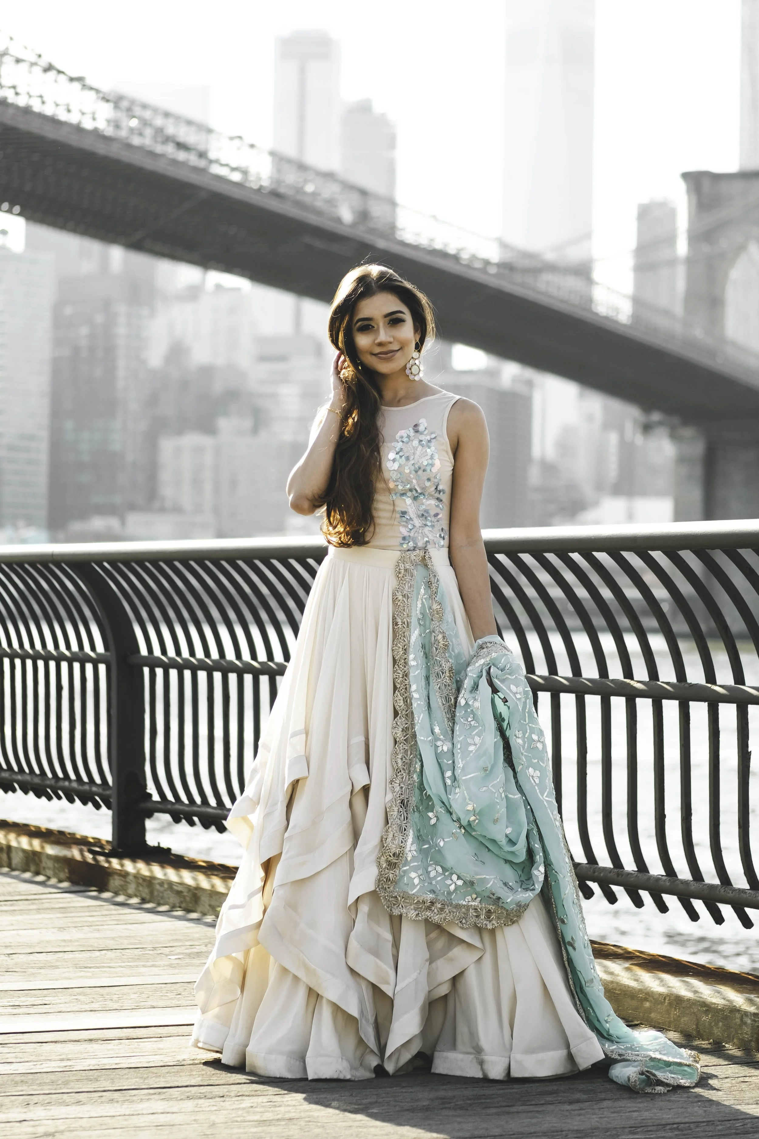 A woman in a white gown with blue accents stands on a boardwalk by the water with a city skyline and bridge in the background.