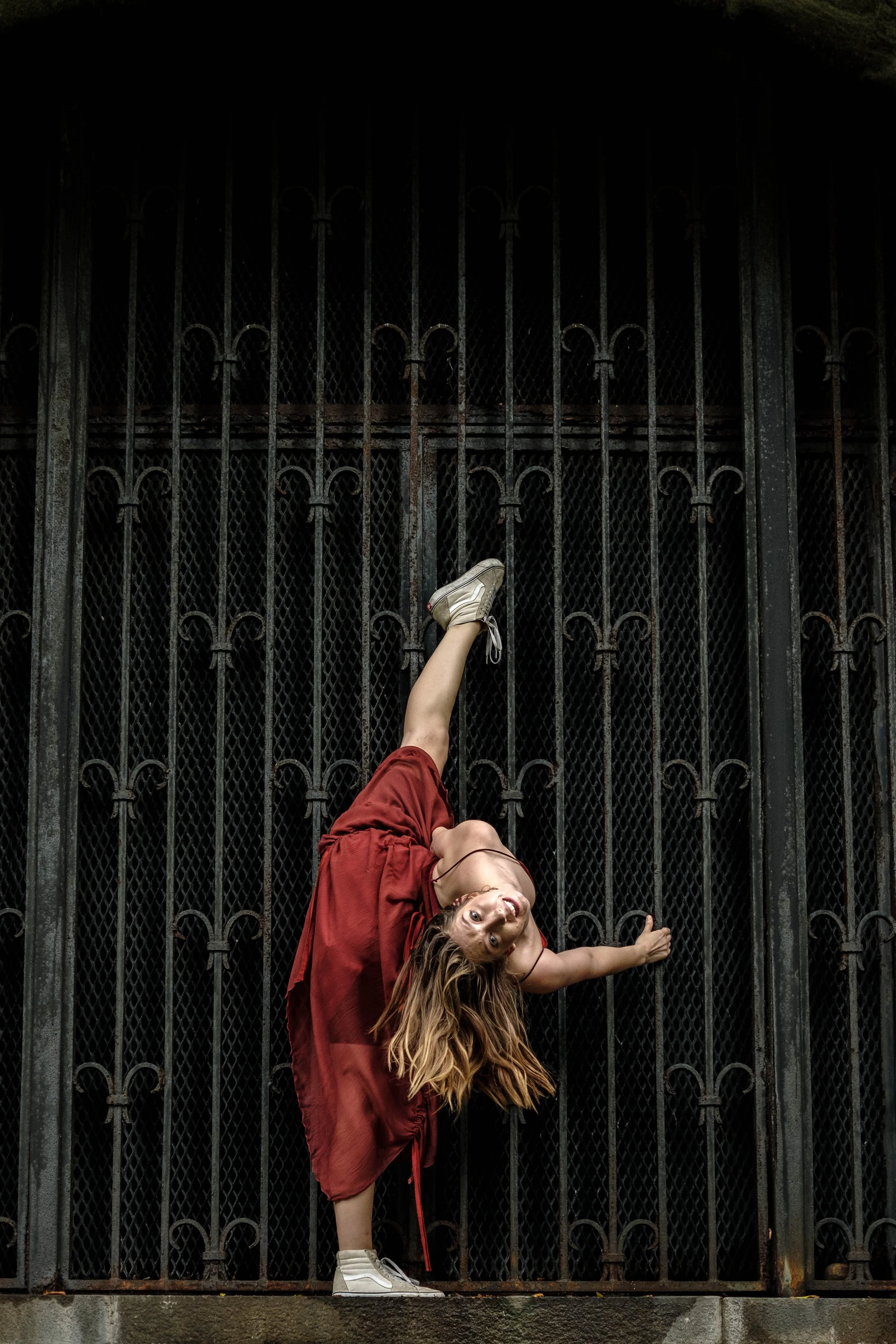 A person with long hair wearing a red jacket and white sneakers is lying on their side, leaning against a large black metal gate with vertical bars and decorative loops, in front of a dark background.
